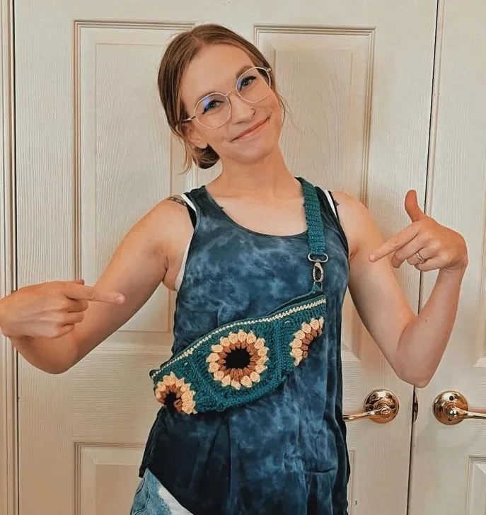 Smiling woman with glasses pointing to her tie-dye tank top and crocheted sunflower-patterned bag, standing in front of a beige door.