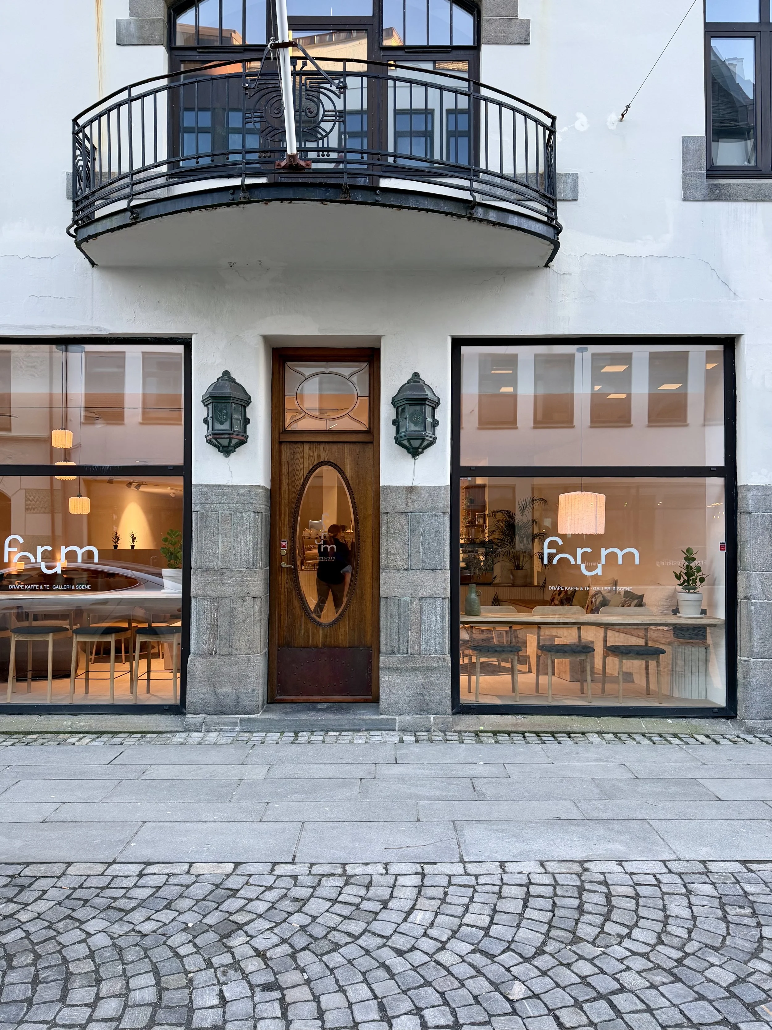 The front of a building with large windows, a wooden door with an oval glass pane, and a small balcony with a black metal railing. The windows display the word 'form' and show the interior lighting and furniture.