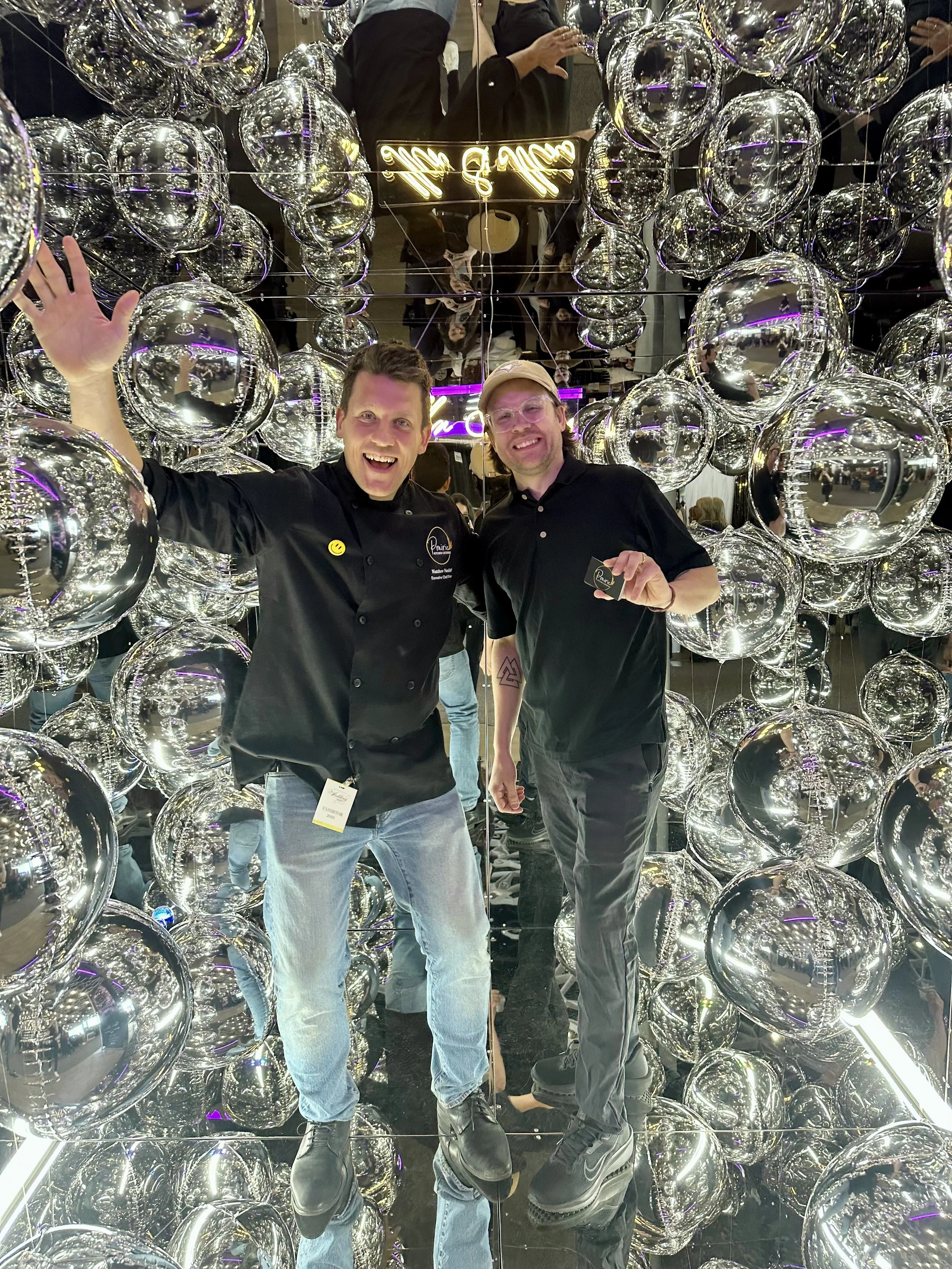 Two men smiling and posing inside a mirror photo booth cube surrounded by reflective silver spheres - foil orbz -, and purple neon lighting.