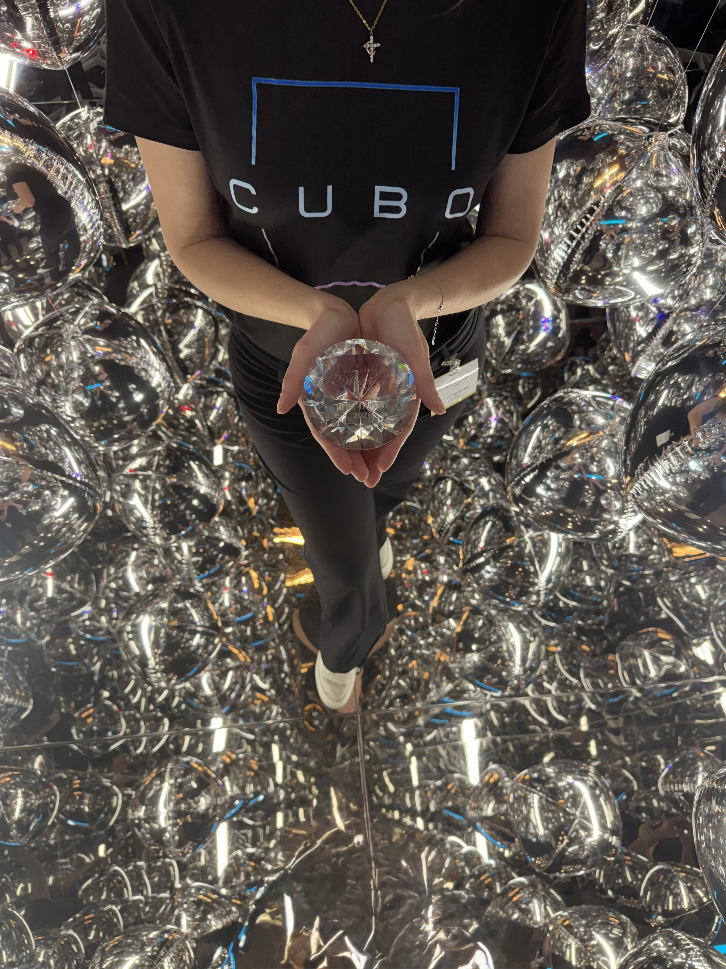Person in black CUBO shirt holding a crystal ornament prize for best wedding show booth while standing in a reflective mirror cube photo booth surrounded by metallic, spherical foil balloons.