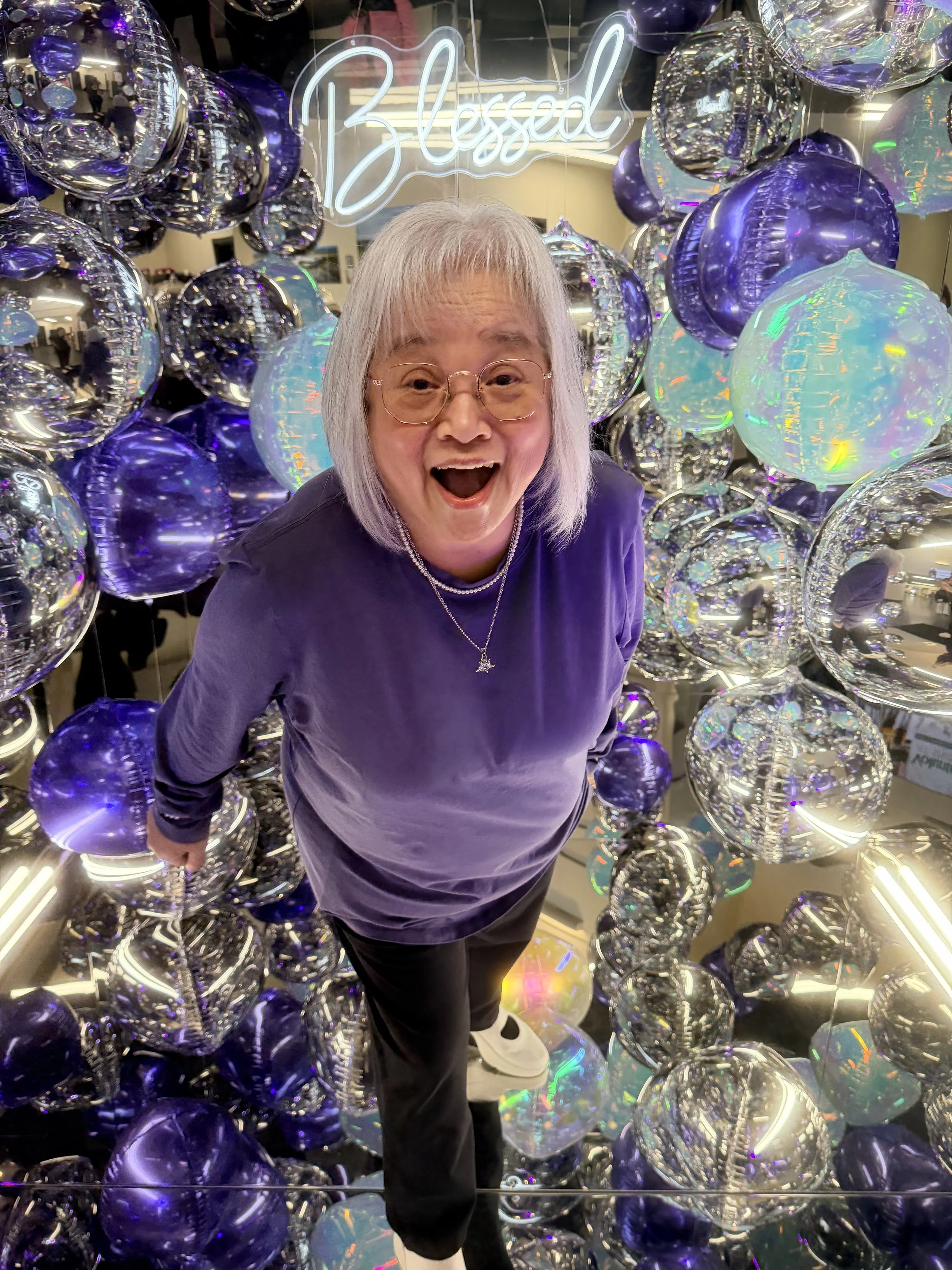 A woman with gray hair, glasses, and a dark purple shirt standing inside a mirror cube photo booth with shiny purple and iridescent balloons, with a neon sign that says 'Blessed' in the background. Conference, corporate.