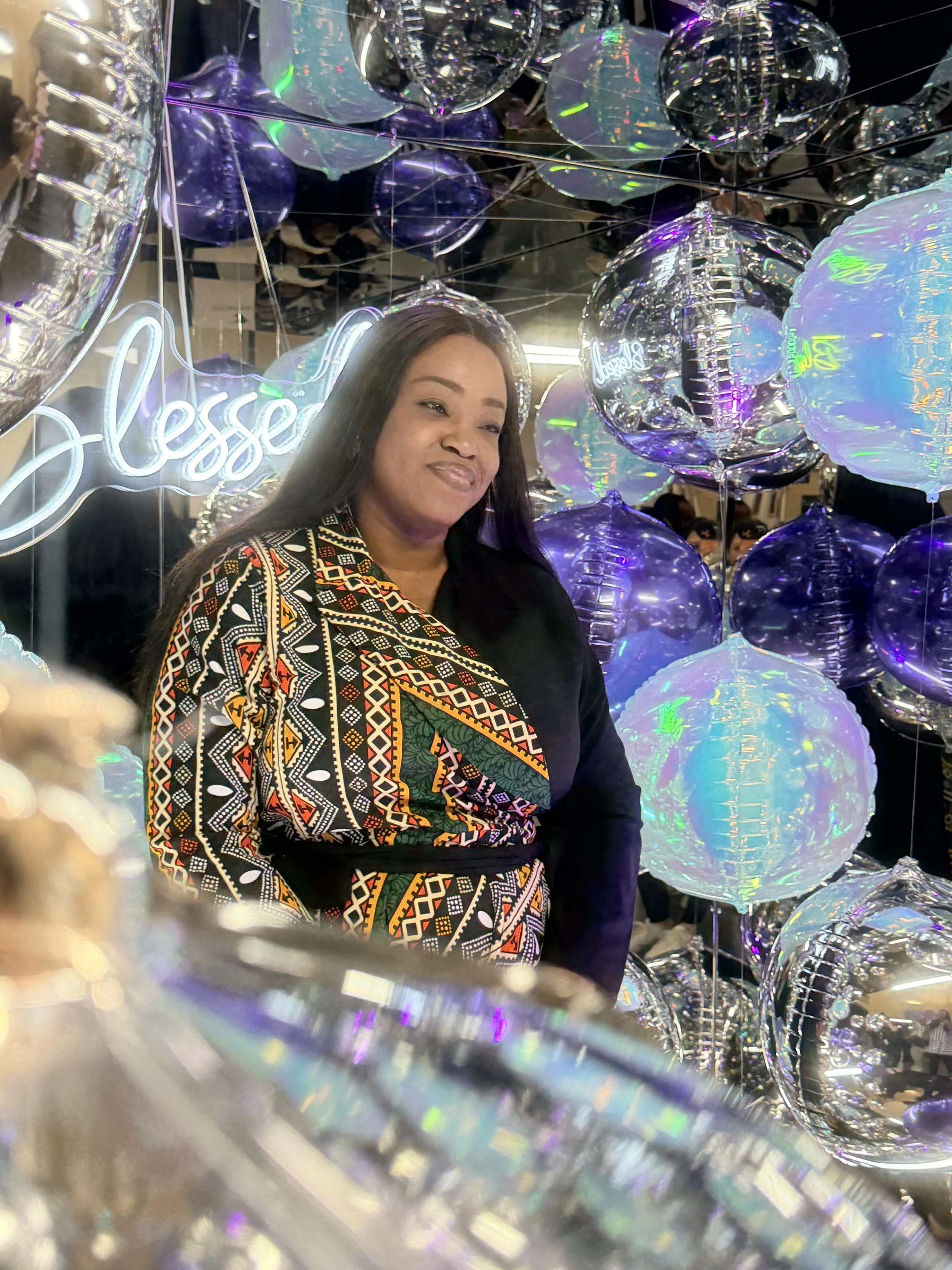 A woman with long dark hair smiling among reflective balloons in various shades of purple and silver, with a neon sign that says 'bless' in the background.