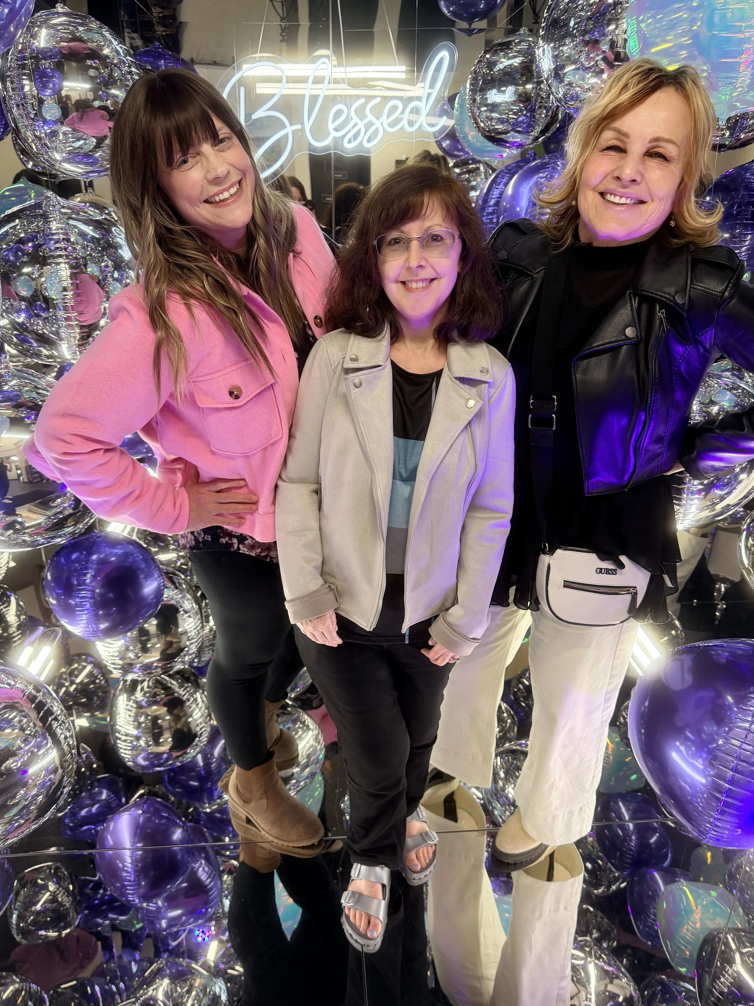 Three women smiling and posing together in a decorated photo booth with shiny purple and holographic balloons. Behind them is a neon sign that says 'Blessed'. This was at a women's conference. They are having fun and are happy and excited.