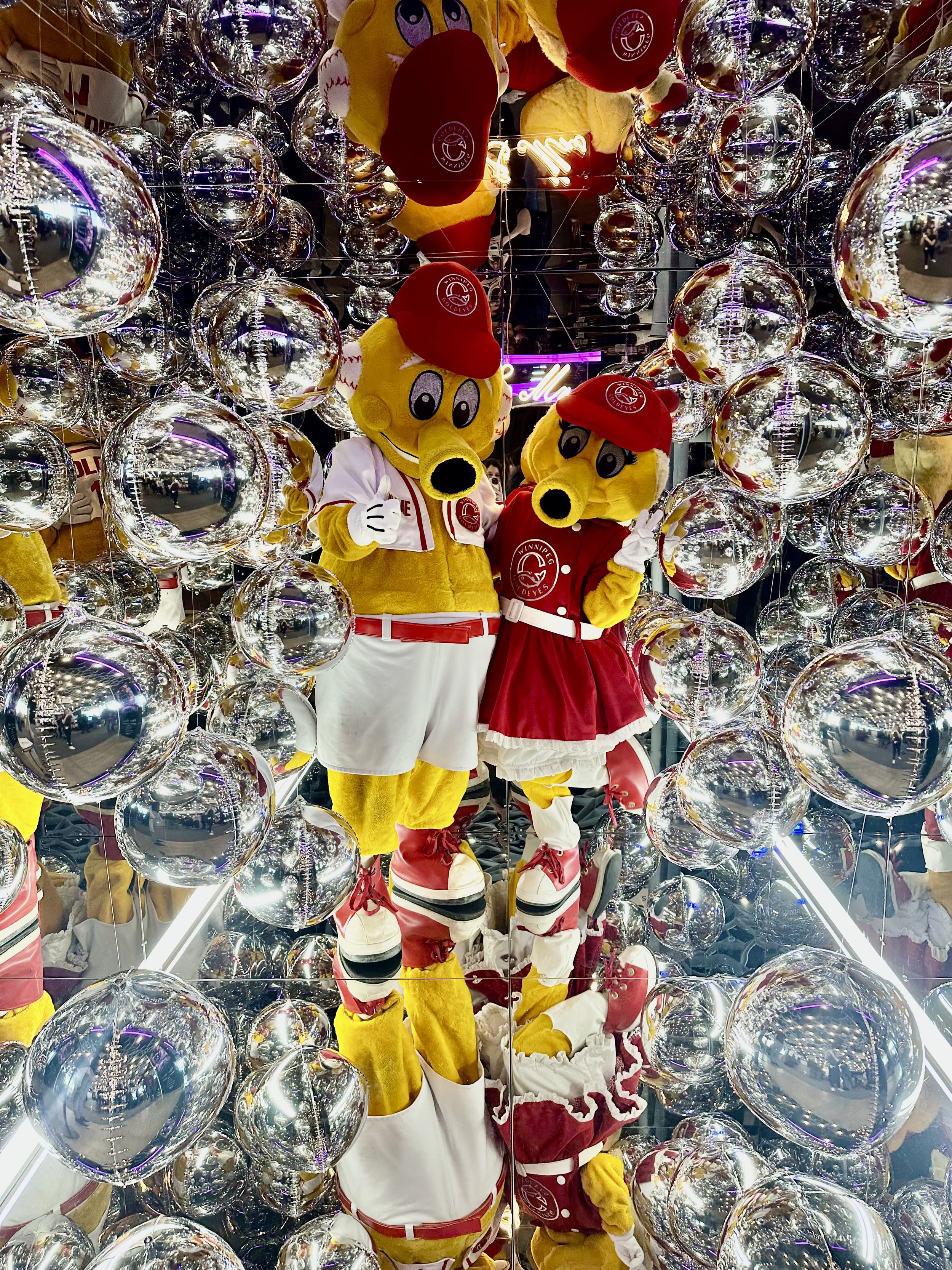 Two mascot characters - Goldie & Goldette from the baseball team 'the Goldeyes' - standing inside a mirror cube phot booth [cabina espejada] surrounded by reflective silver baloons.