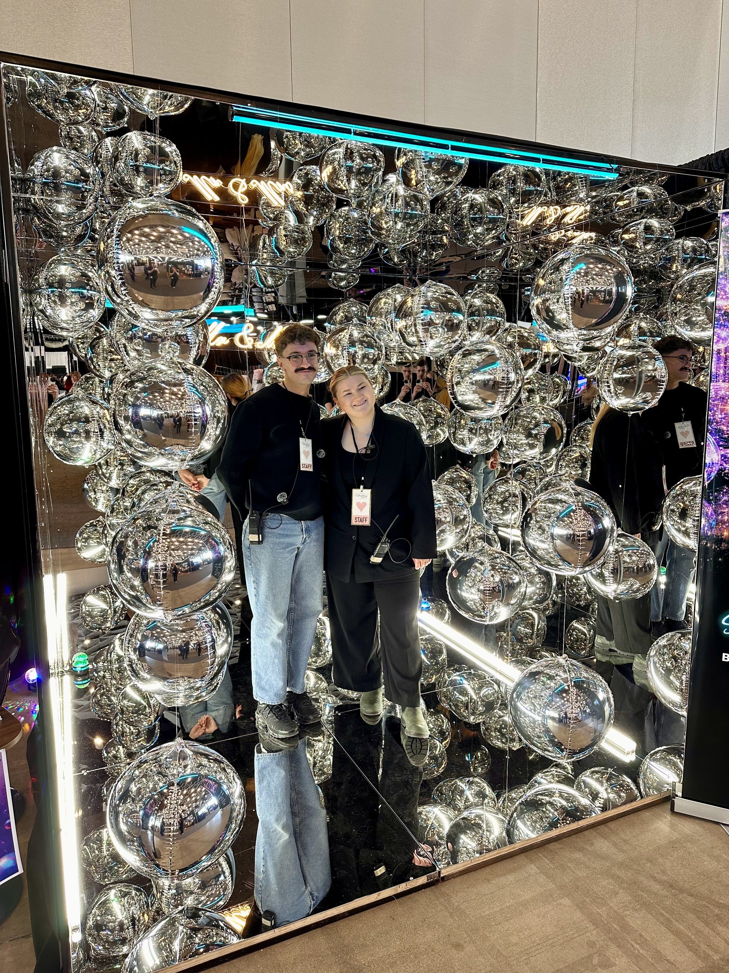 Two people standing in front of a mirror photo booth with silver spheres, orbz, shiny balls and neon lights, smiling for a photo at an event or wedding, corporate event, party, graduation, convocation, gala, fundraiser, bar mitzvah, quince, birthday.