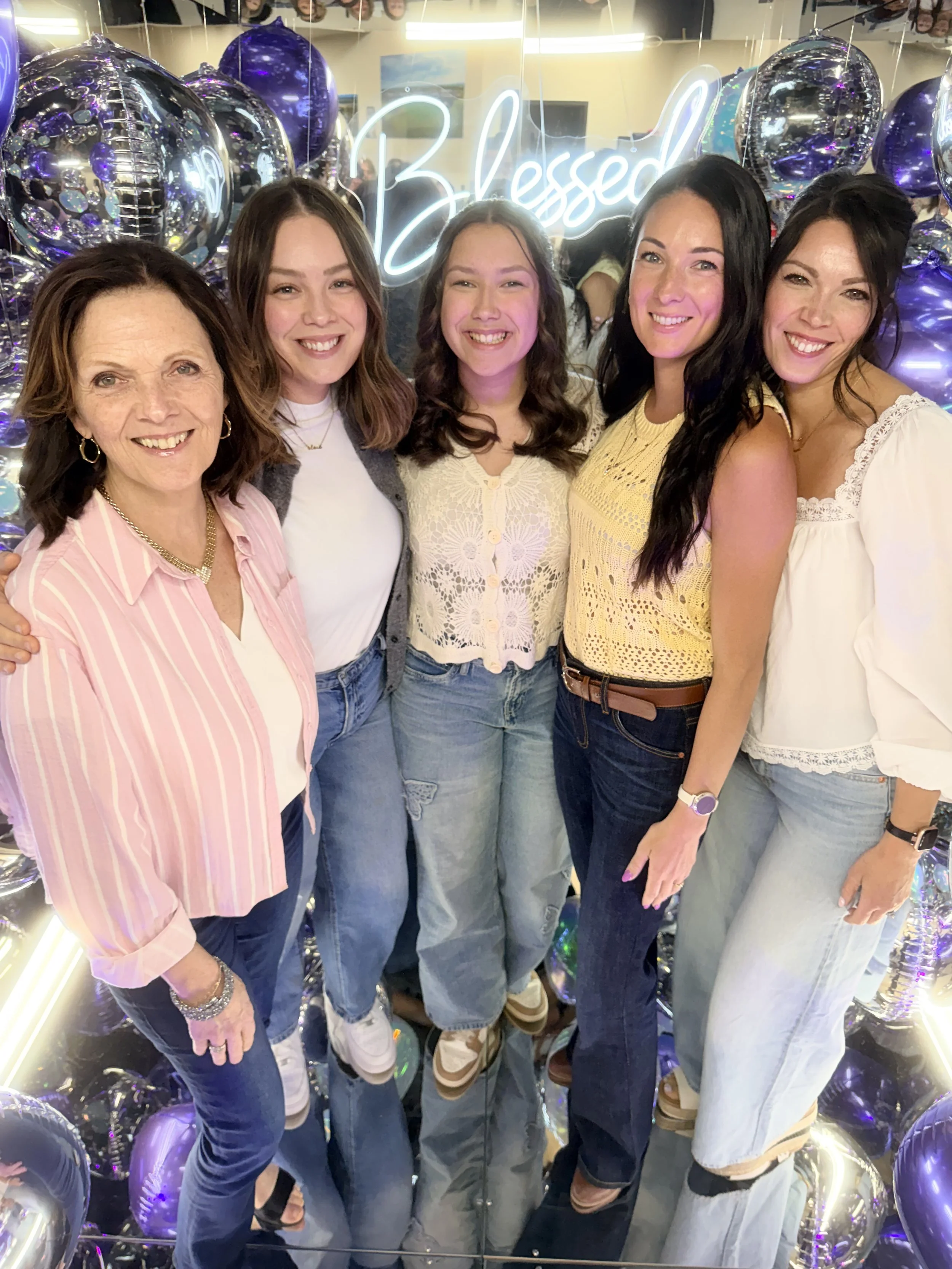 Mom and daughters standing close together in a mirror photo booth with a neon sign that reads 'Blessed' and surrounded by silver and purple balloons, smiling at the camera. having fun. celebrating. celebration