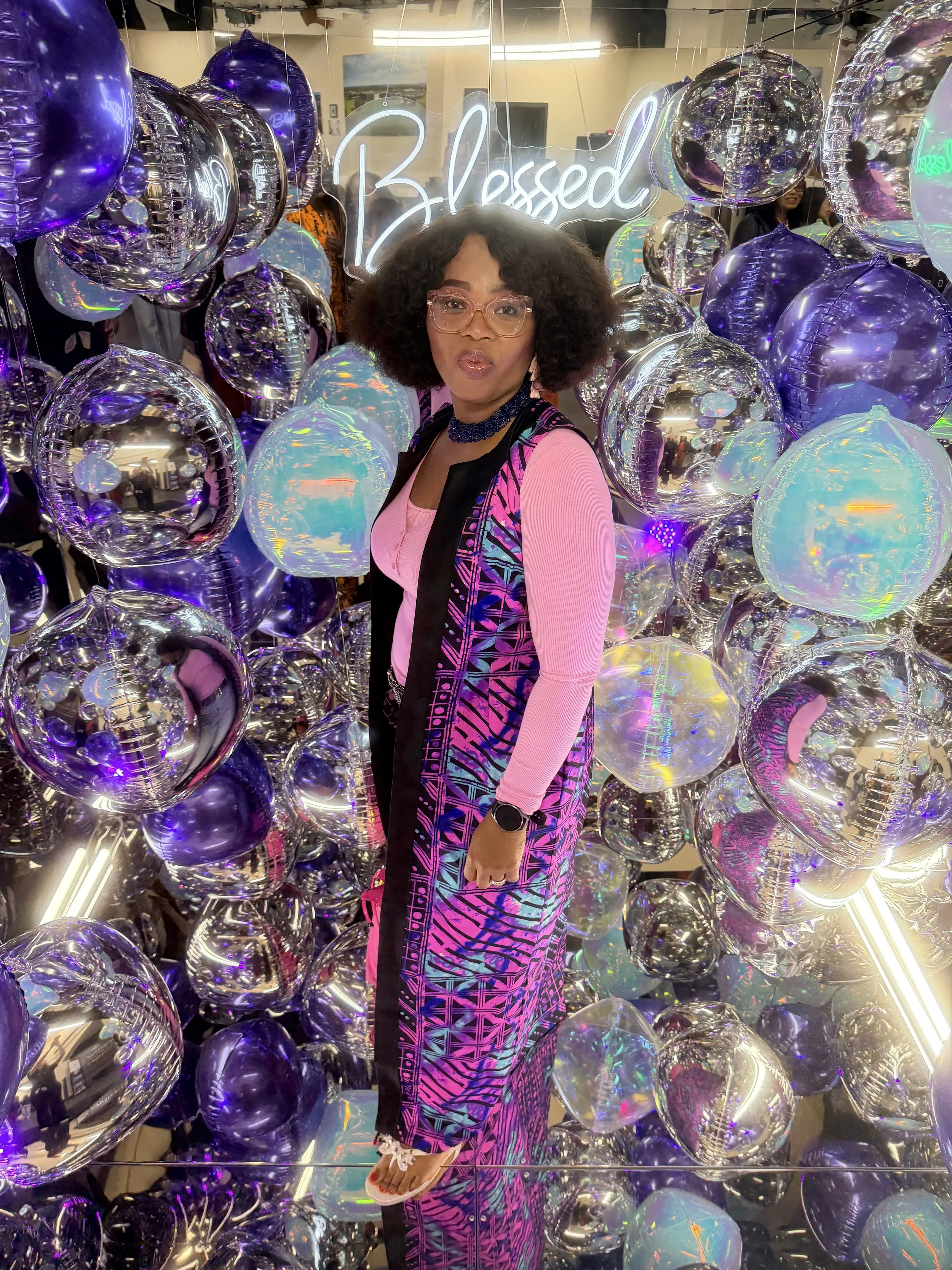 A woman with curly hair, wearing glasses, and a colorful patterned outfit, standing among metallic purple and iridescent balloons in a photo booth. Behind her, a neon sign reads 'Blessed'. Corporate, celebration, brand activation, conference