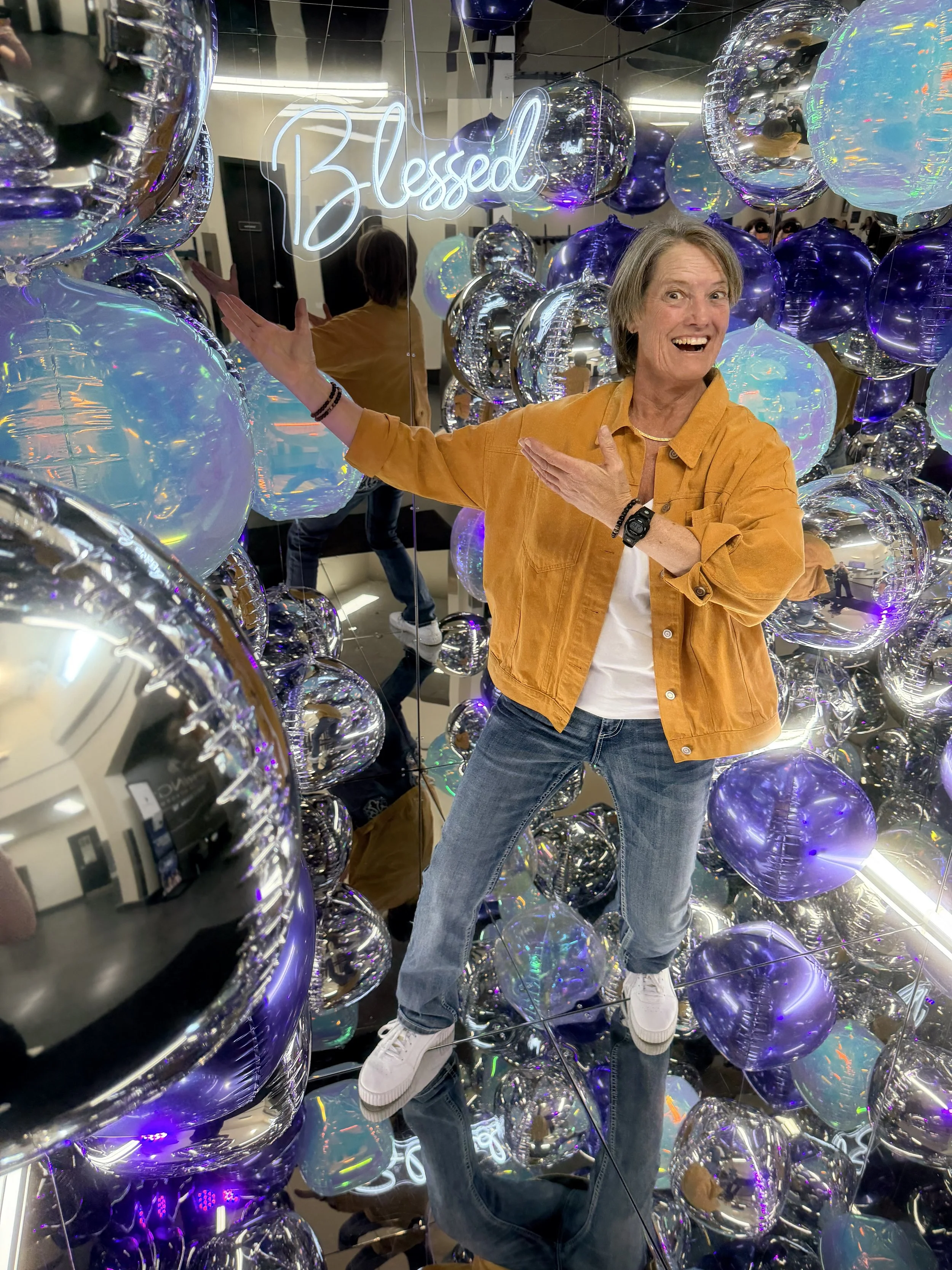 A woman smiling and posing while surrounded by numerous purple and iridescent balloons in a mirror cube photo booth with a neon sign that says 'Blessed' in the background.