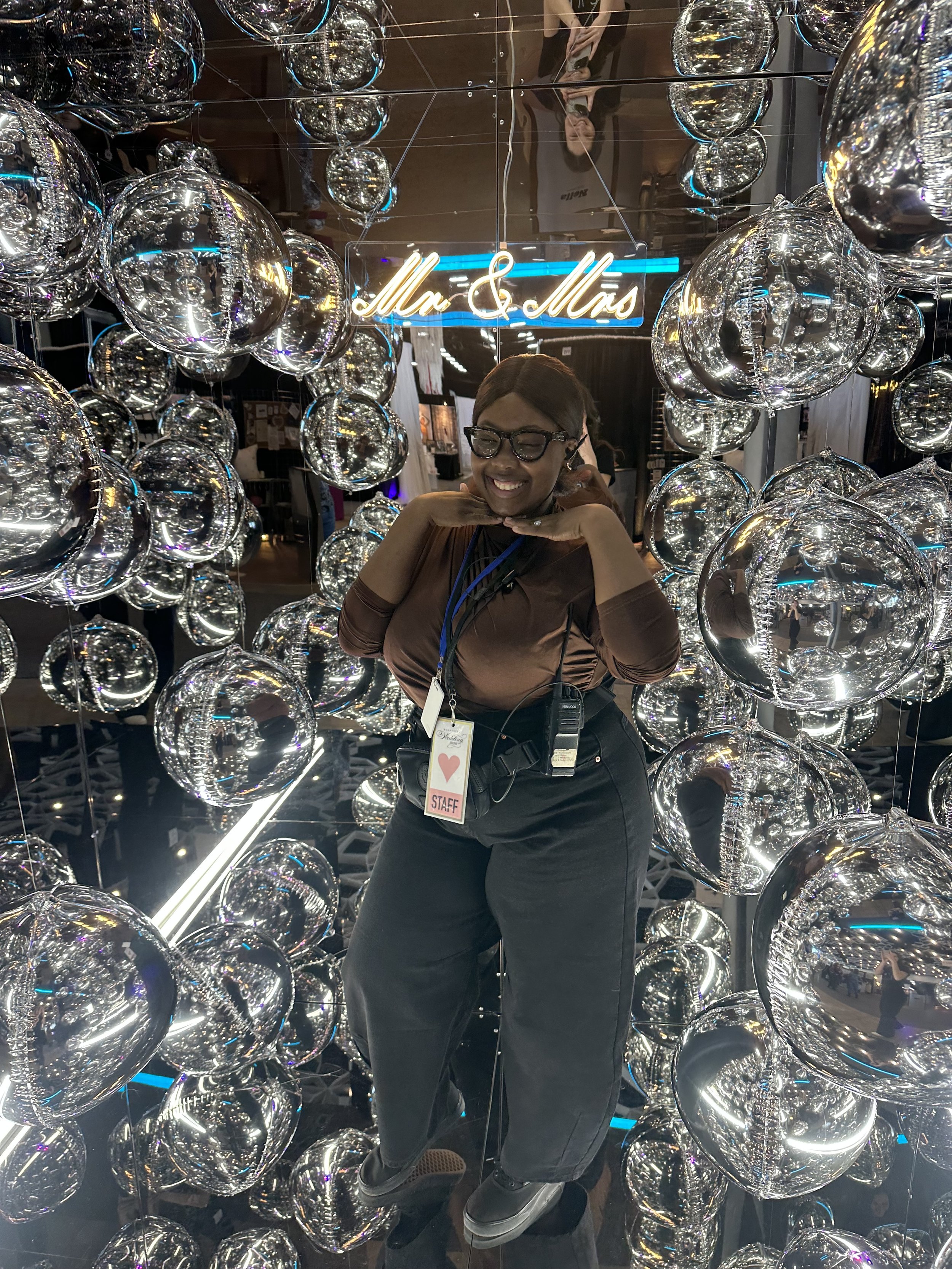 Woman smiling inside a mirrored cube photo booth decorated with numerous silver foil balloons, with a neon sign reading 'Mr & Mrs' in the background.