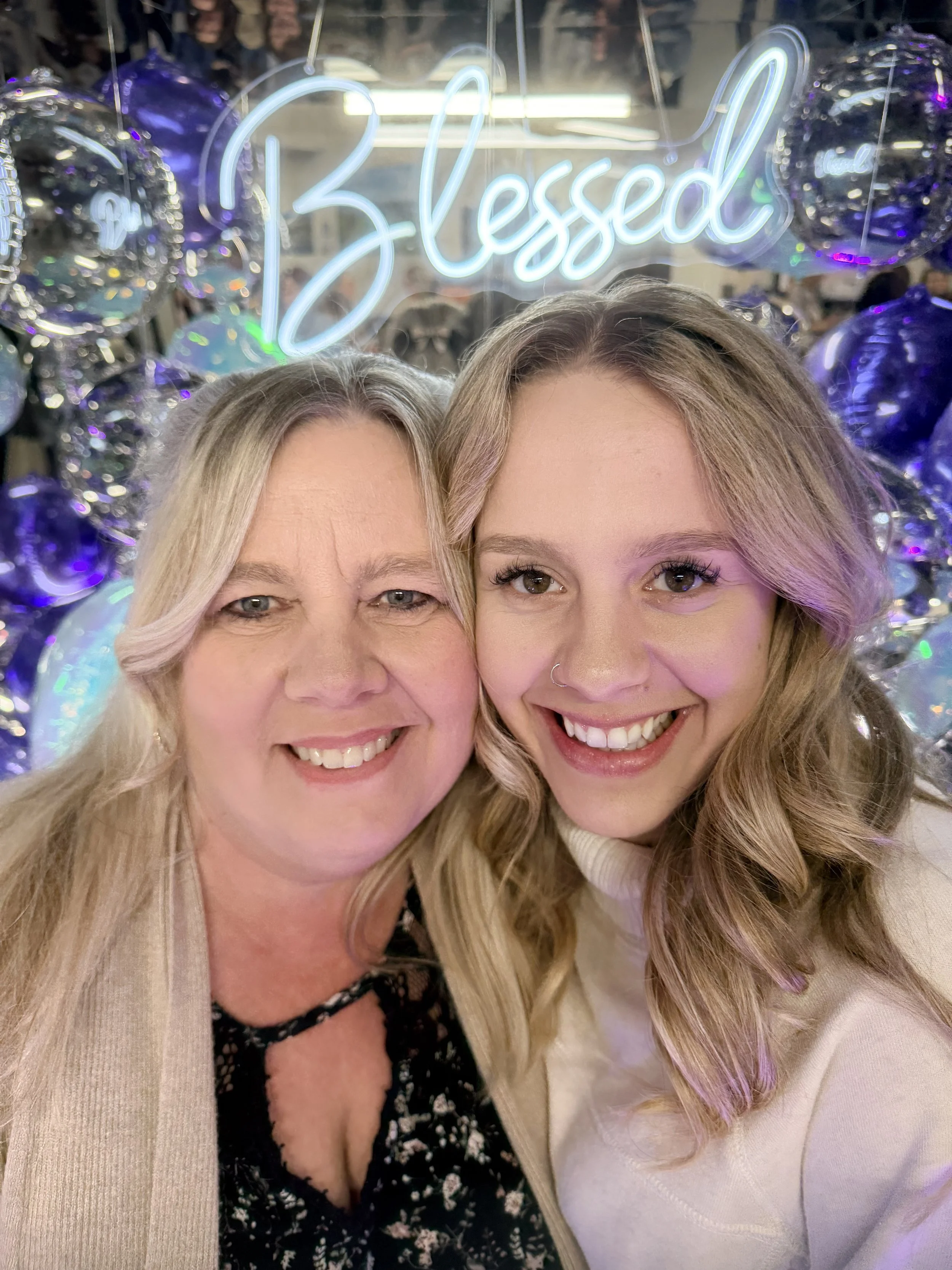 Two women smiling in front of a backlit sign that says 'Blessed,' with metallic balloons and festive decorations behind them. They are in an experience photo booth at a conference.