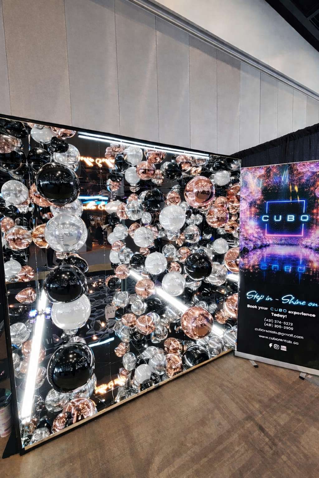 Mirrored cube photo booth with black, white, and rose gold shiny balloons and a sign promoting a space called CUBO for booking experiences at a event centre for weddings, corporate events, parties, graduations, convocations, galas, fundraisers.