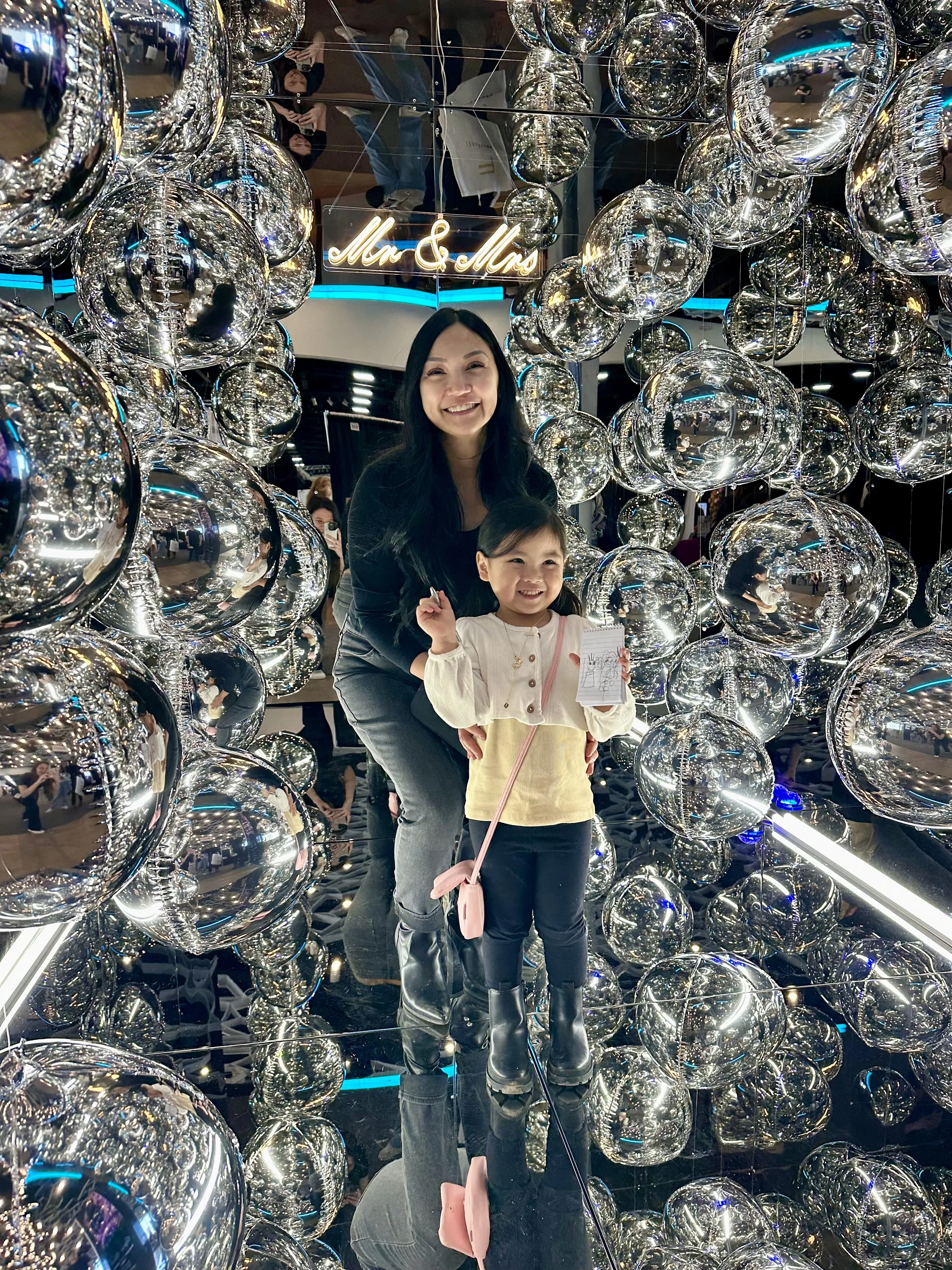 A woman and a young girl smiling inside a reflective, mirrored structure booth decorated with shiny, metallic balls, orbz. The girl holds a small notebook and a pink bag, and a sign that says "Love". In the background, a neon sign reads "Mr & Mrs".