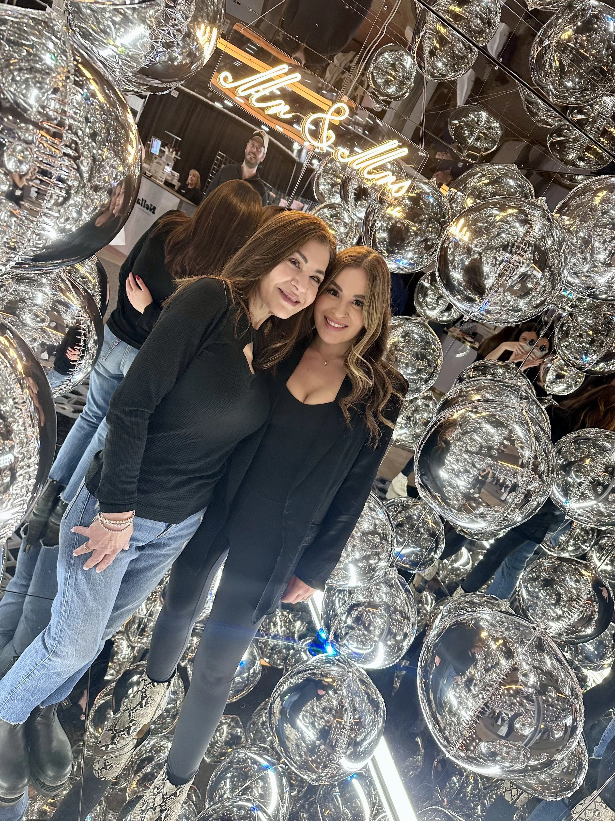 Two women standing together, smiling at the camera, surrounded by shiny silver balloons and mirrors at an event or exhibition with a neon sign in the background that reads 'Mr & Mrs'.