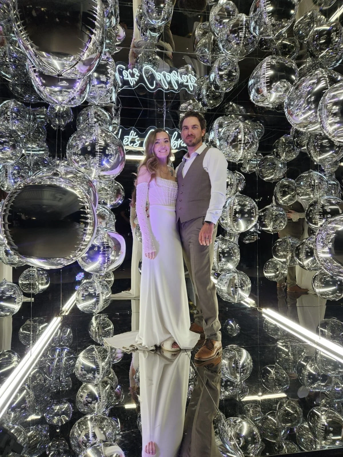 A bride and groom standing in a display of hanging silver balloons and mirrors, with a neon sign in the background that says 'Let's Celebrate'.