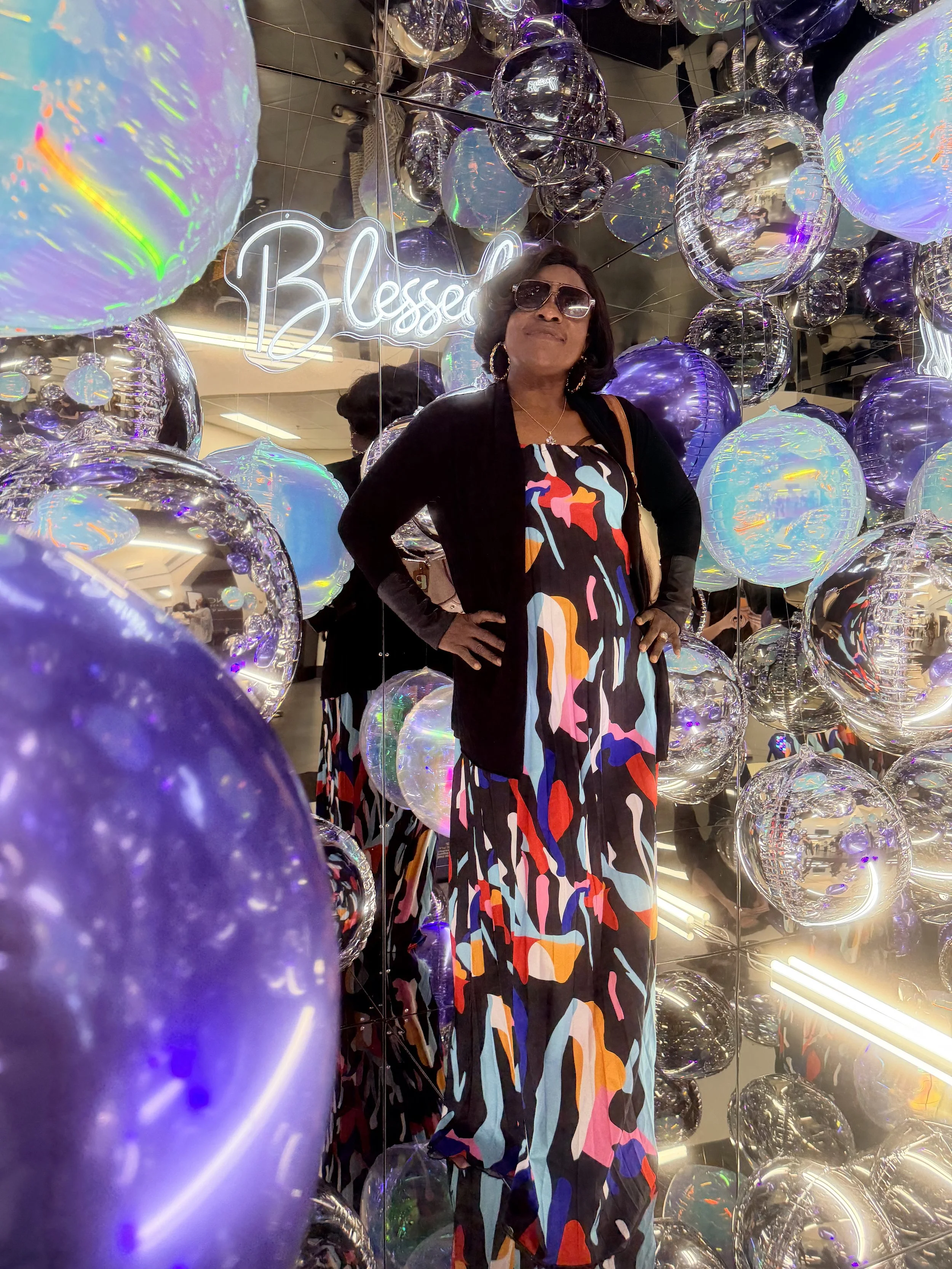 A fashionable woman wearing sunglasses and earrings, standing inside a mirror cube photo booth among reflective purple and silver balloons, with a neon sign that says 'Blessed' in the background at a women's conference.
