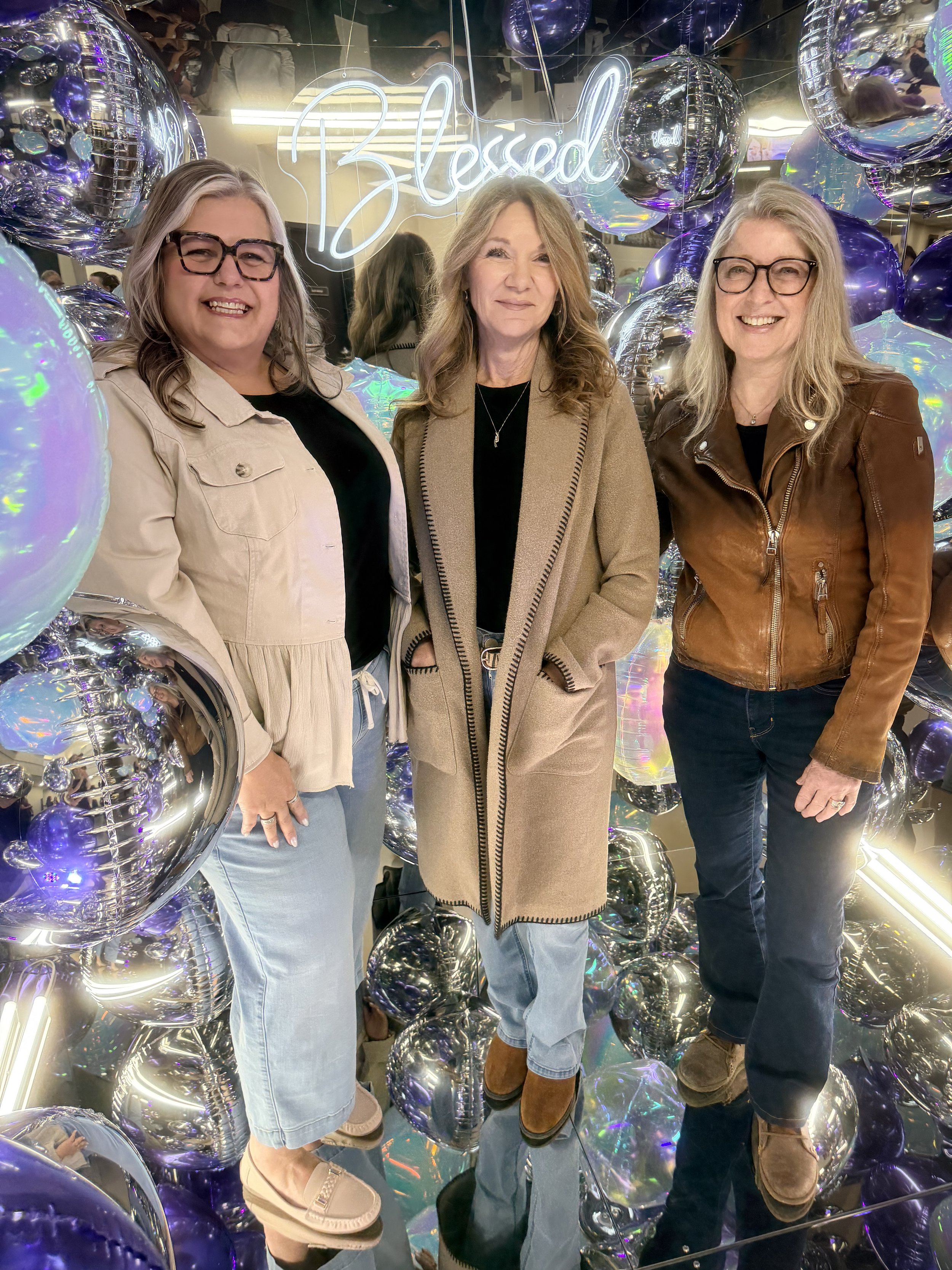 Three women standing inside of a mirror photo booth surrounded by silver and iridescent balloons. Behind them is a neon sign that reads 'Blessed'. The women are smiling and posing for the photo, dressed casually and wearing glasses.