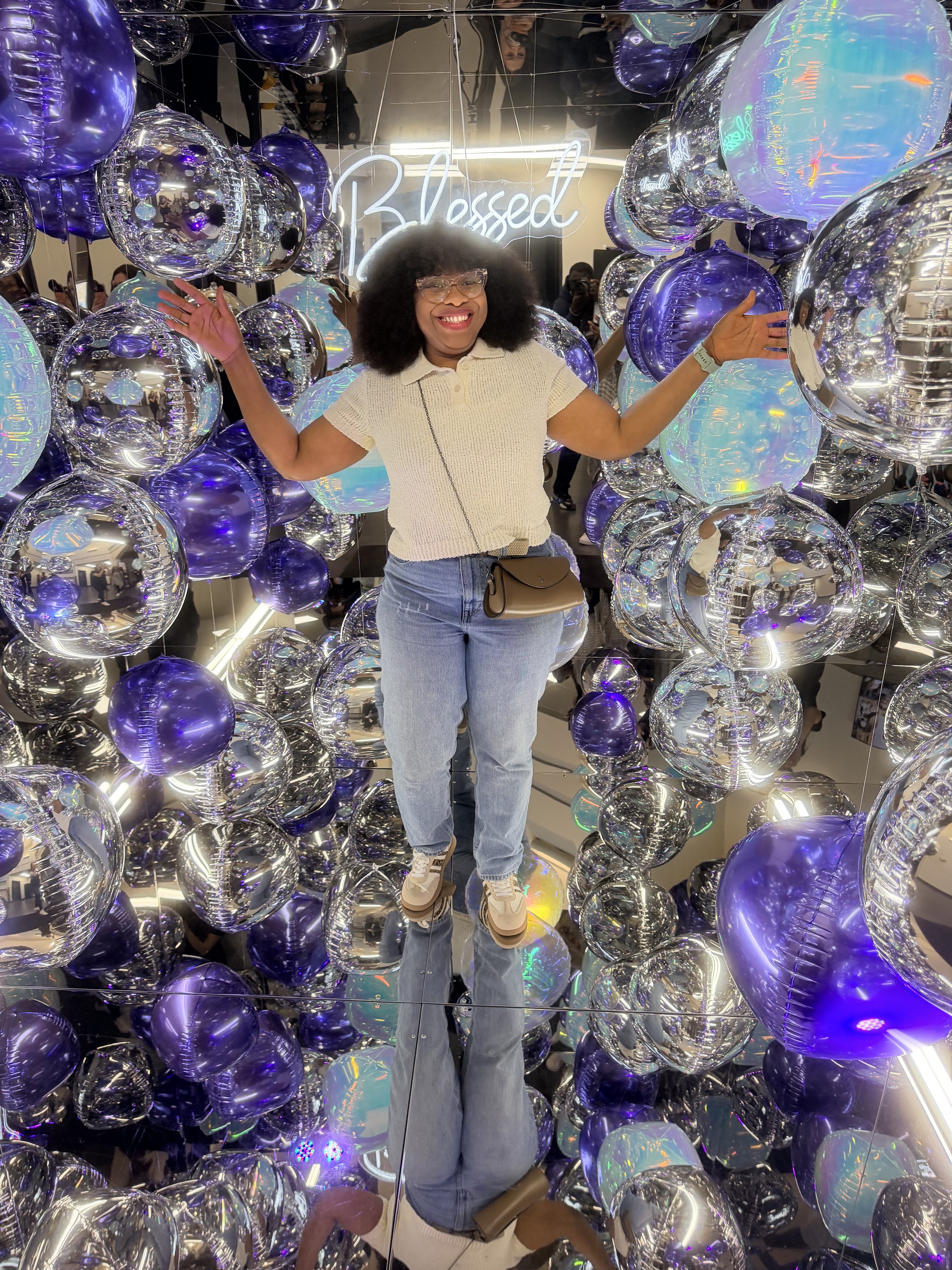 A woman with curly hair and glasses standing amid purple and iridescent balloons inside of a mirror cube photo booth with a neon sign that says 'Blessed' at a women's conference. The woman is having fun, is joyful and excited.