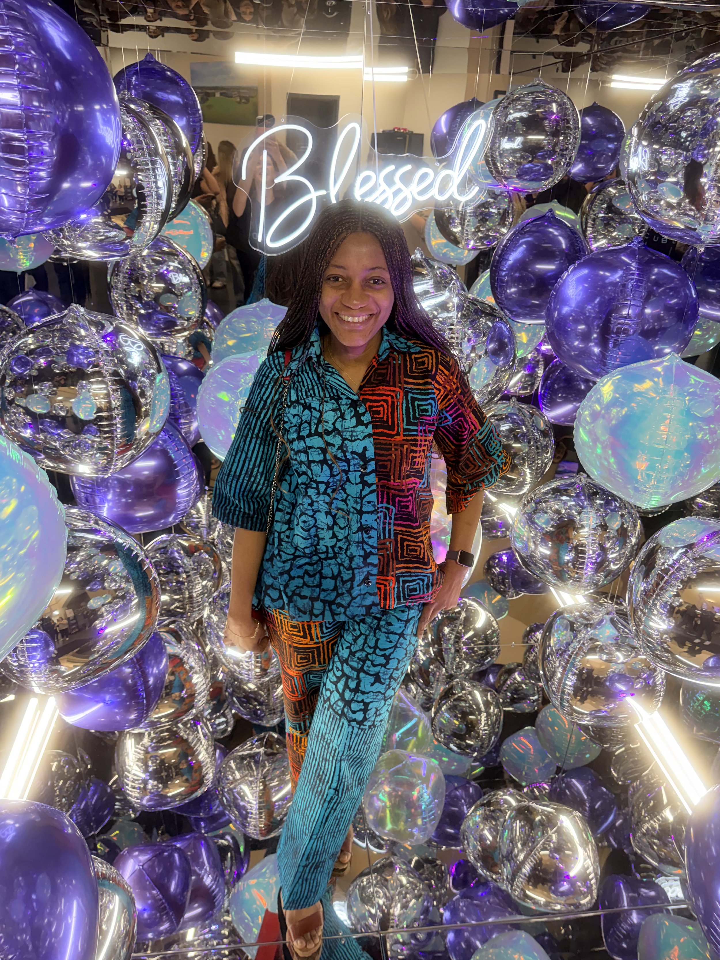 A smiling woman standing among a large number of purple, silver, and iridescent balloons, with a neon sign that reads 'Blessed' behind her inside a mirror cube photo booth.