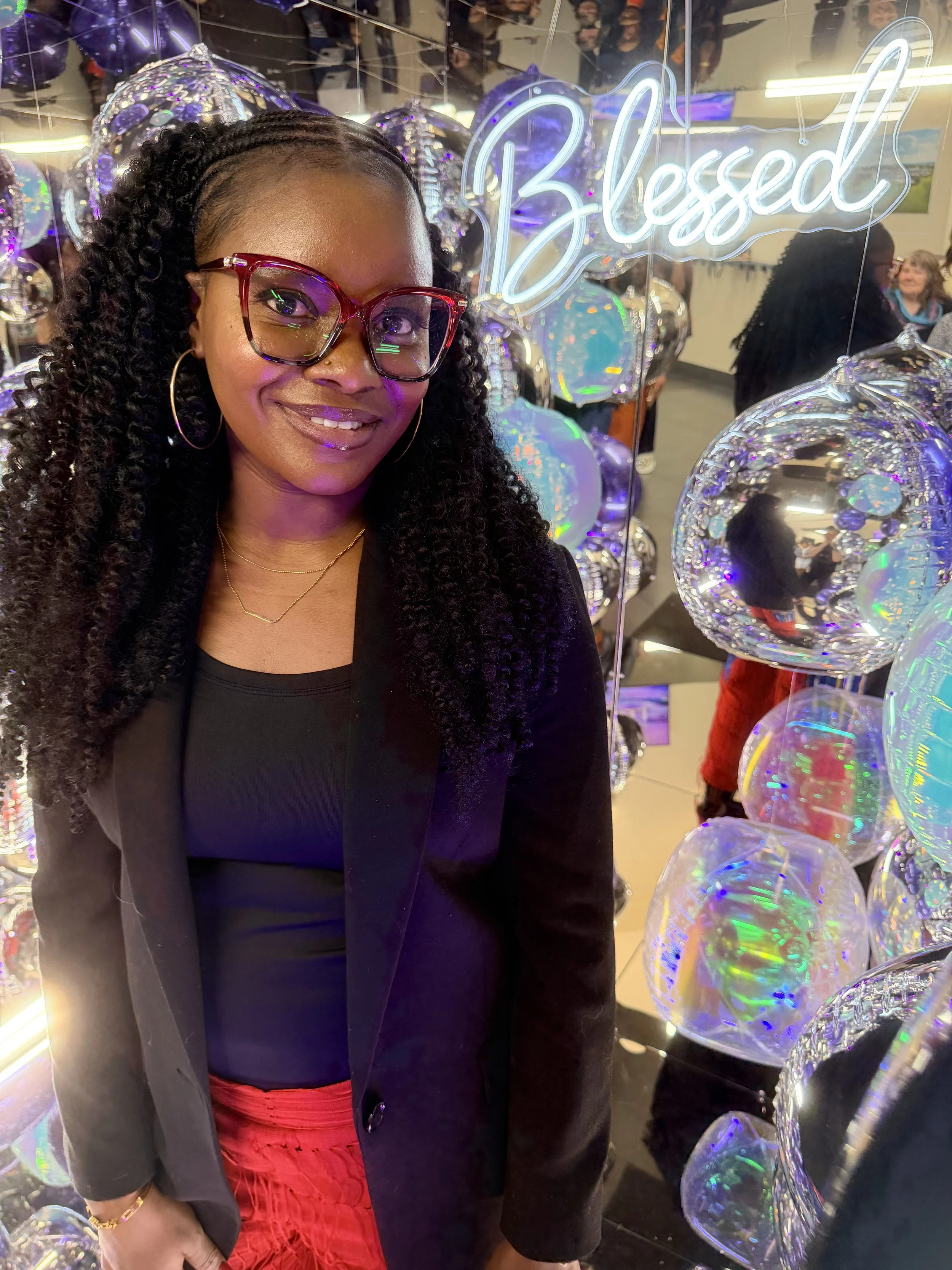 A woman with curly hair, glasses, and hoop earrings smiling in a  mirror cube photo booth surrounded by orbs and reflective balloons and a neon sign that says 'Blessed'. Brand activation, conference, corporate.