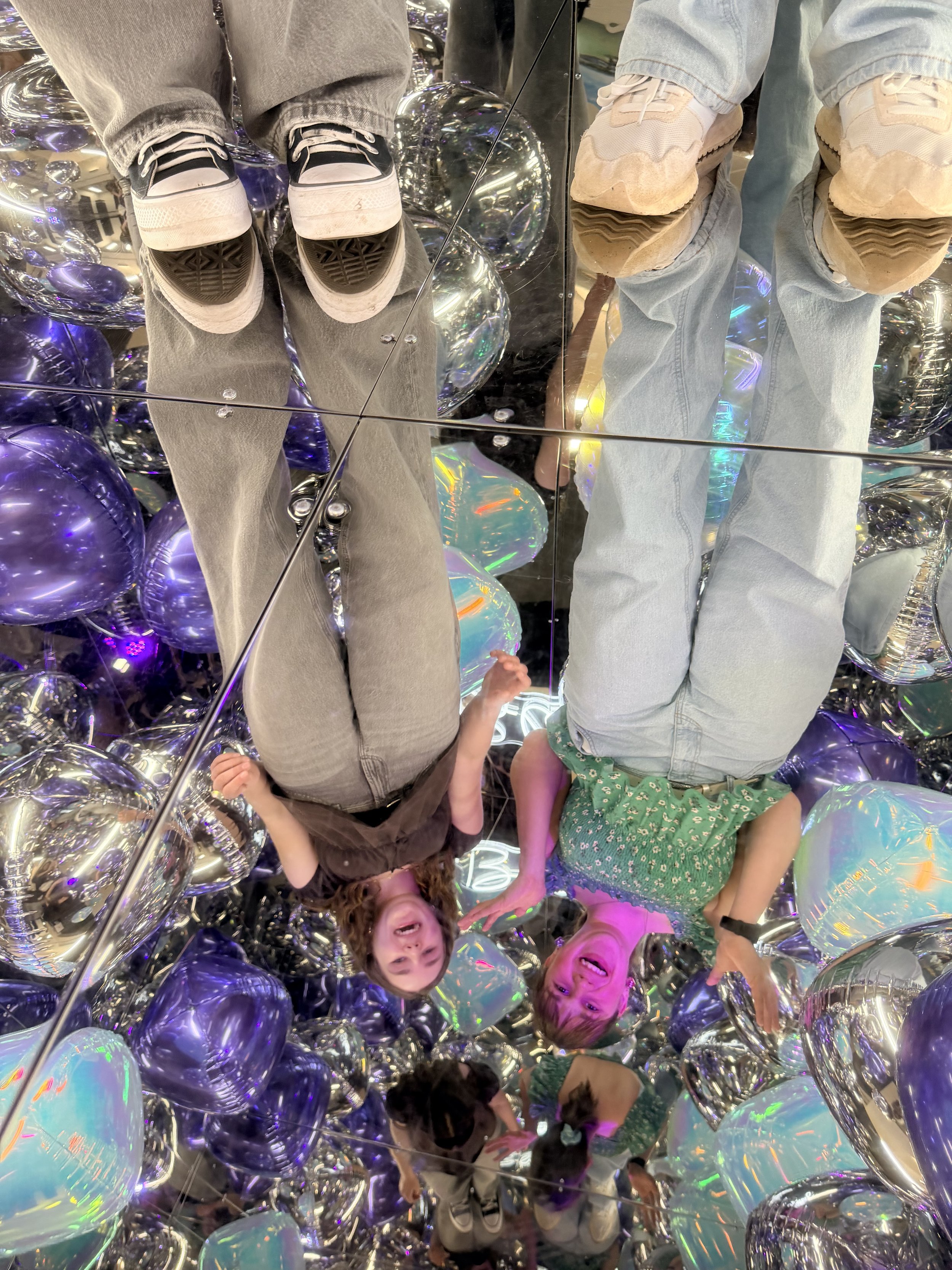 People standing on a reflective surface inside a mirror photo booth surrounded by shiny silver and purple balloons, with their reflections visible in a mirror. unique picture, having fun, celebrating.