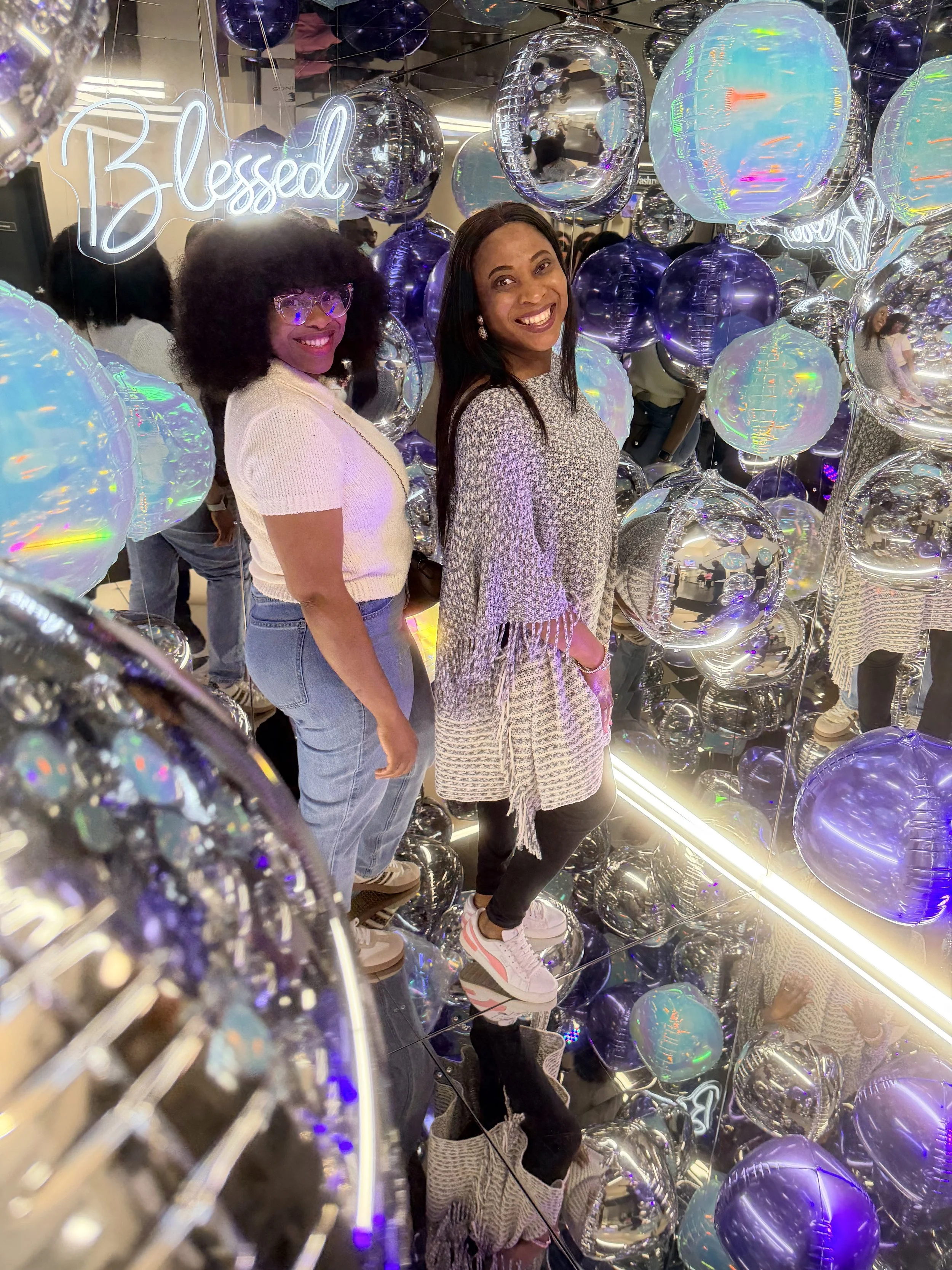 Two women smiling inside a mirror photo booth surrounded by numerous colorful balloons, including a neon sign that reads 'Blessed' in the background having fun at a conference.