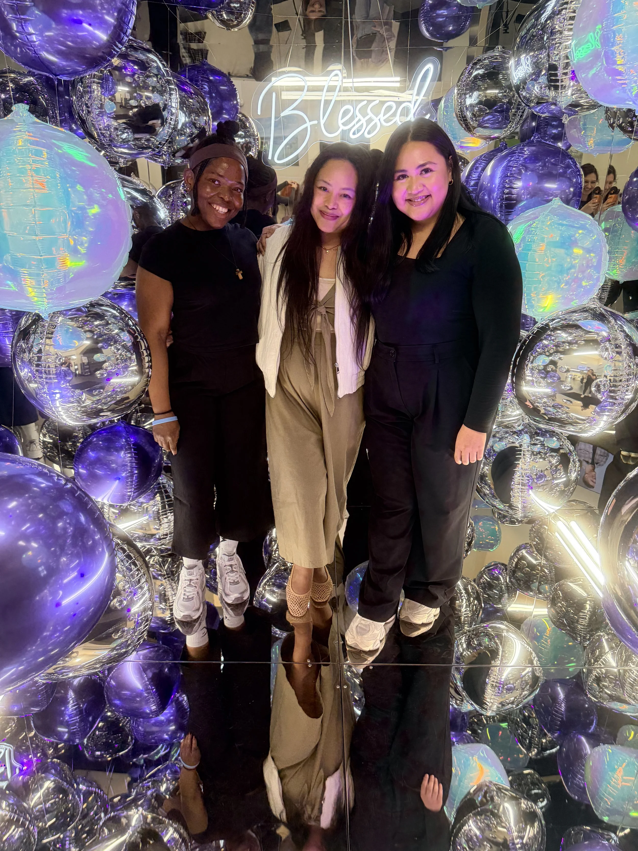 Three women standing together surrounded by metallic purple and iridescent balloons in front of a neon 'Blessed' sign at a celebration or party. Photo booth.