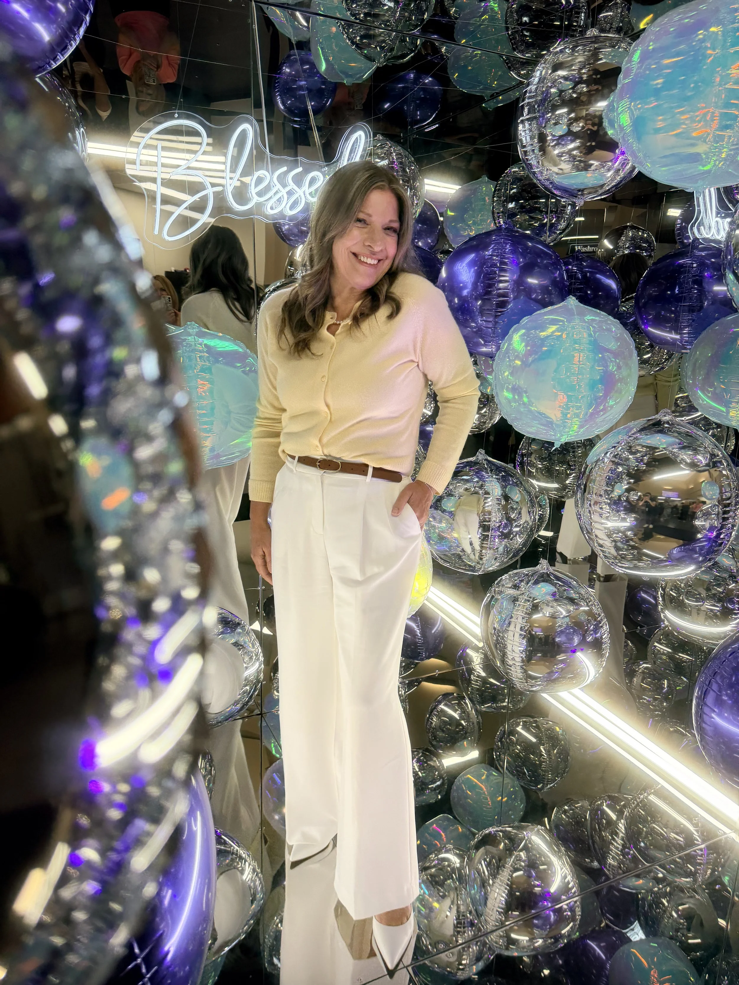 A woman smiling and posing among shiny, reflective balloons in various shades of purple and iridescent colors. She is inside an illuminated decor photo booth with a neon sign that says 'Blessed' and mirrors reflecting other people and the space.