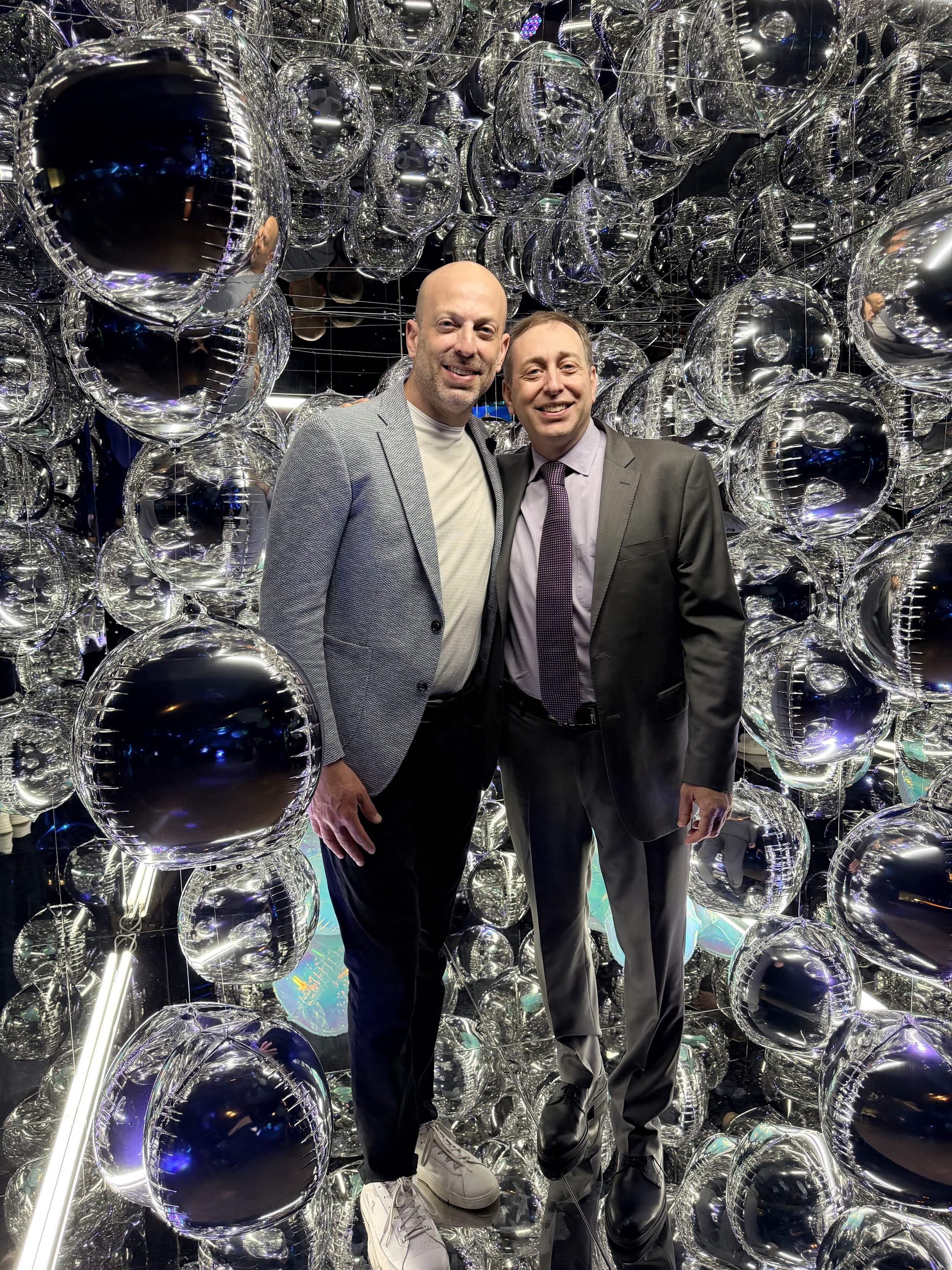 two men in suits at a bar mitzvah smiling and looking at the camera at an angle inside the mirror photo cube booth