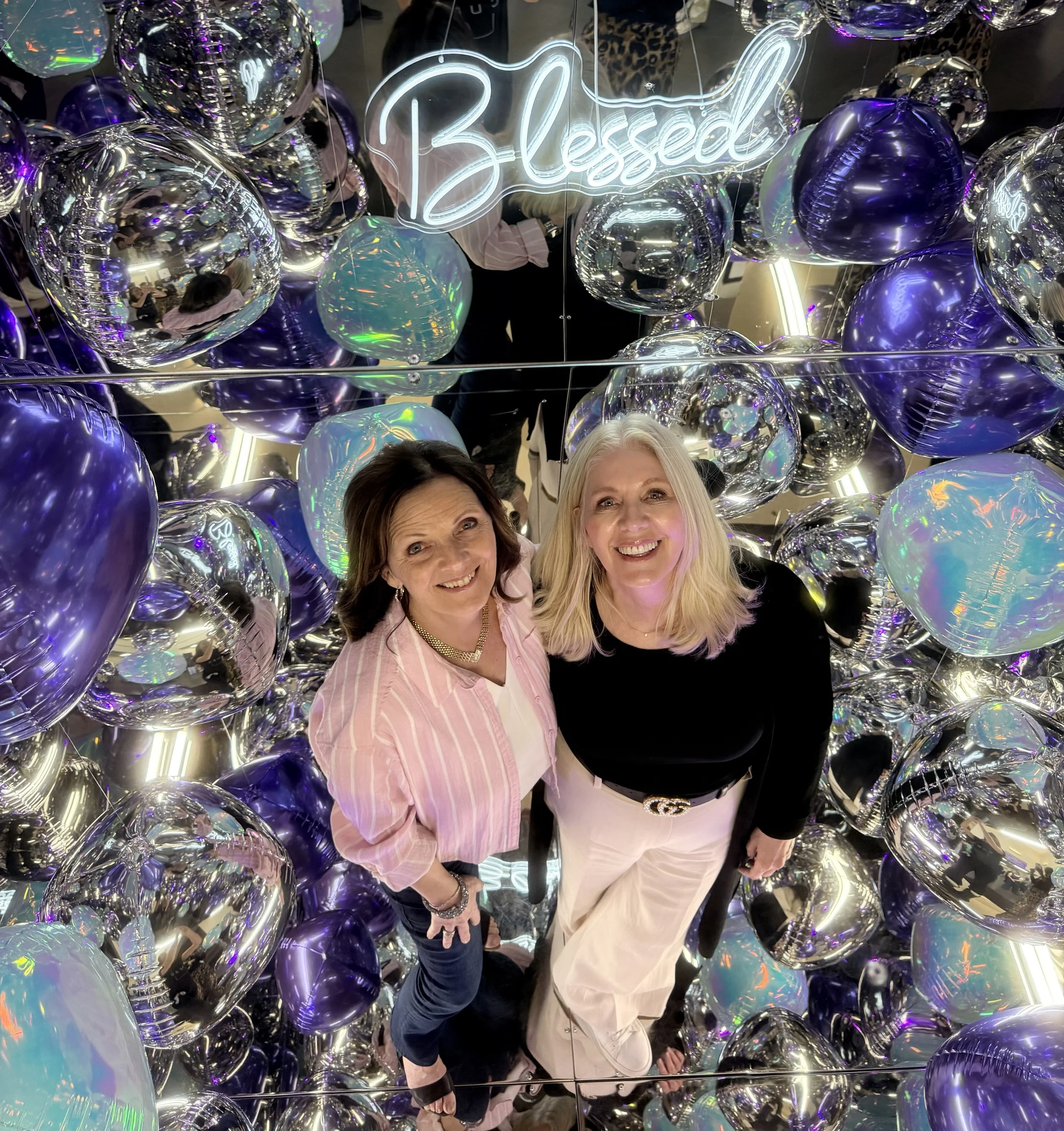 Two women smiling among a colorful display of reflective balloons and neon signs with the word "Blessed" in cursive.