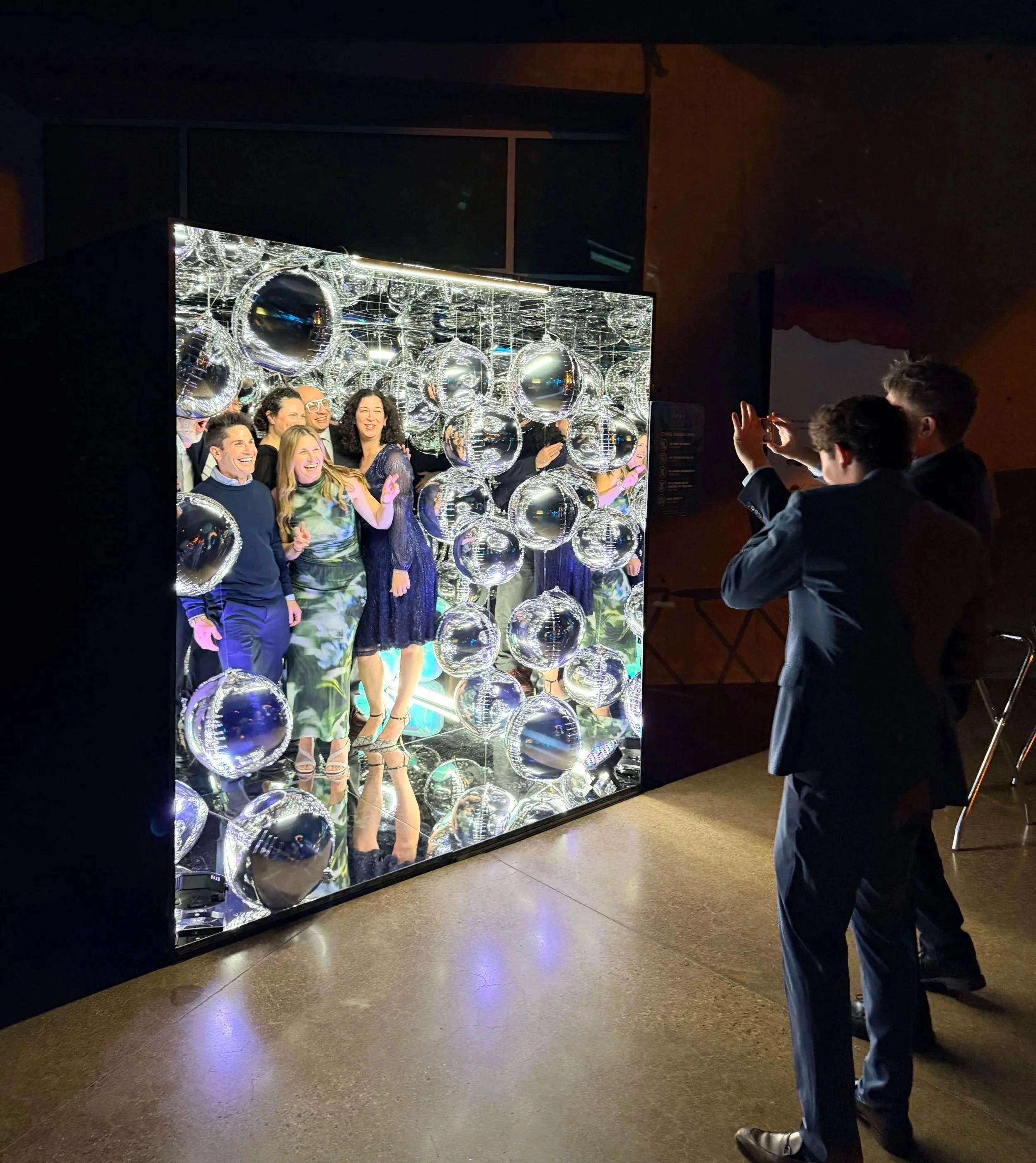 People posing for a photo inside a mirror booth decorated with shiny metallic spheres, with a person taking their picture on a smartphone.