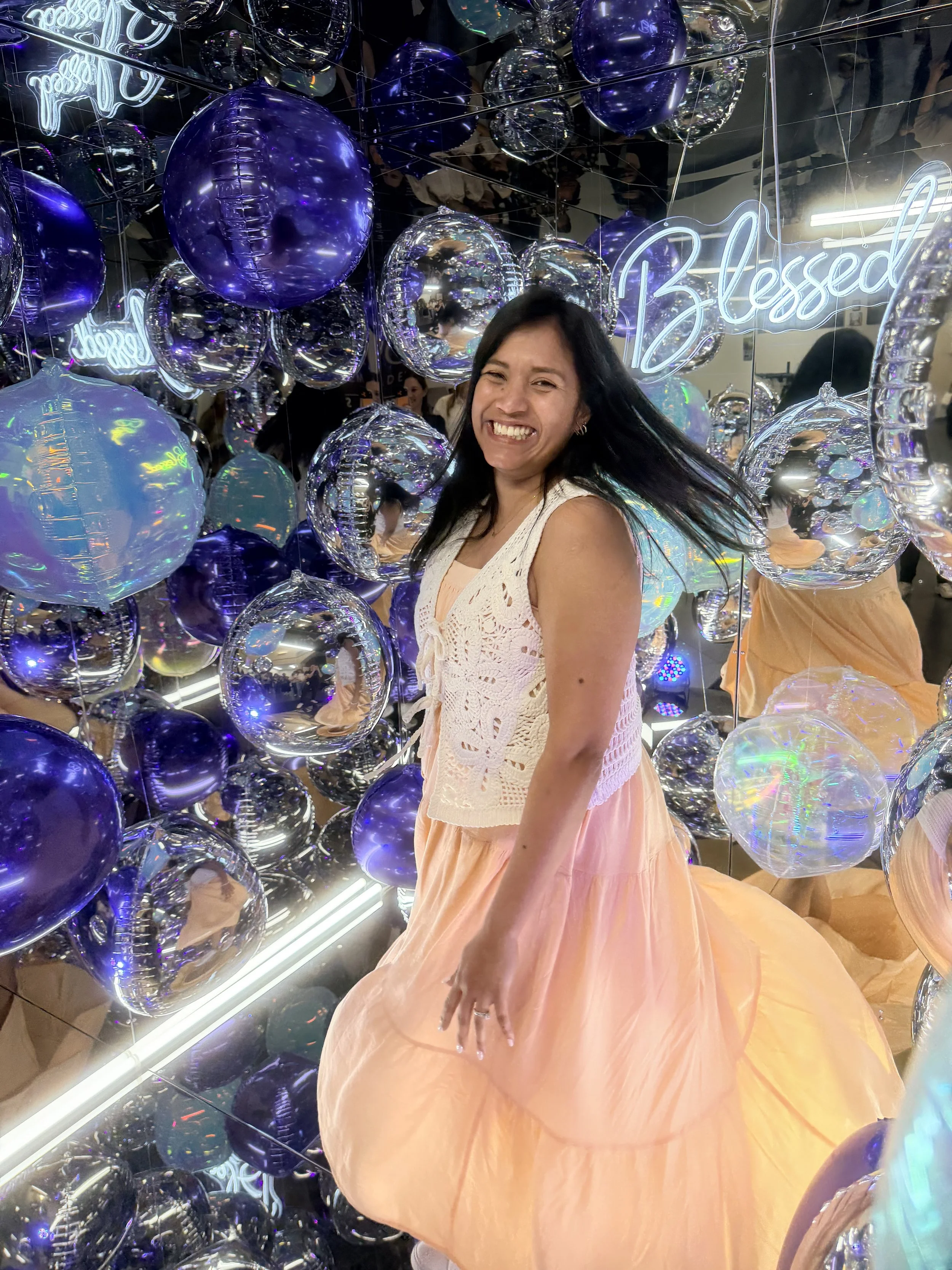 A woman with black hair and a big smile poses and is having fun inside a mirror photo booth among numerous purple and clear balloons in front of a neon sign that says 'Blessed.'