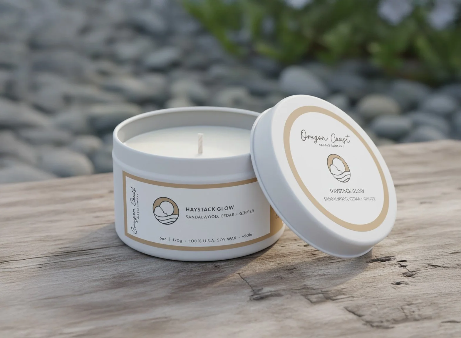 A white scented candle in a round container with beige and white labels resting on a wooden surface outdoors. The candle is labeled "Oregon Coast" and "Haystack Glow," with scents of sandalwood, cedar, and ginger. The candle's lid is leaning against the container.