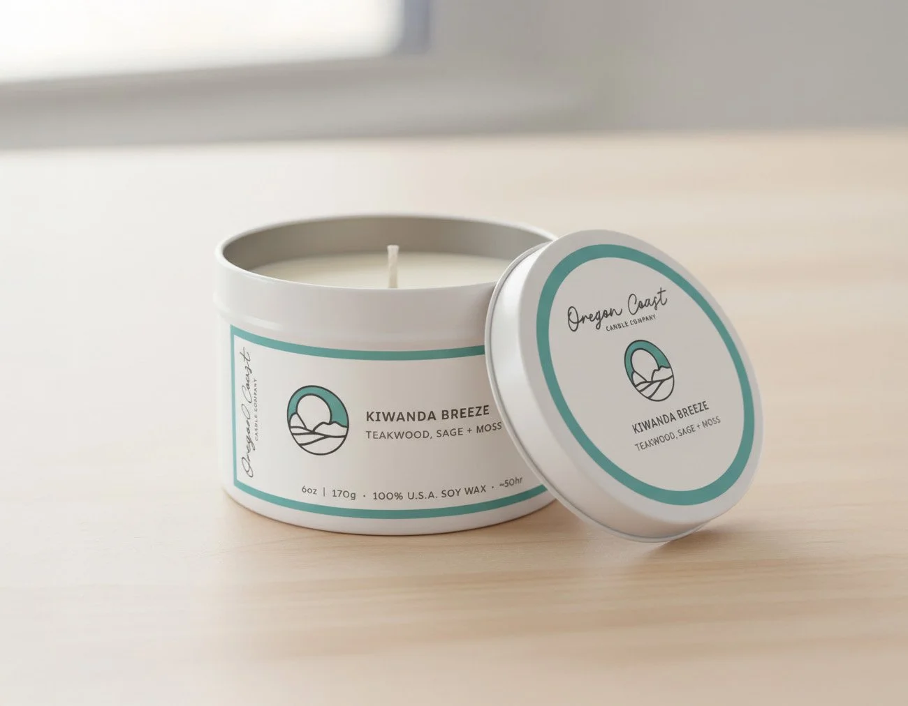 A white scented candle in a round container with a removable lid, labeled 'Oregon Coast Candle Company' and 'Kiwana Breeze' with scents like teakwood, sage, and moss, on a light wooden surface.