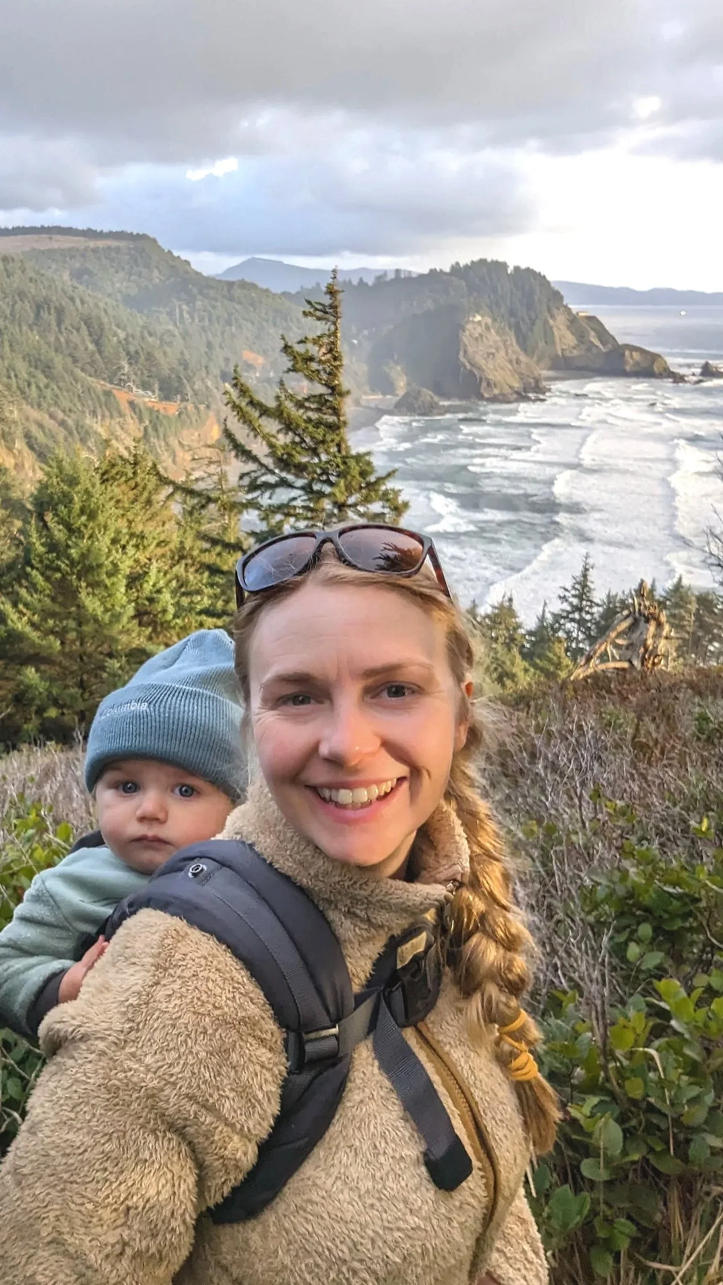 A woman smiling while carrying a baby on her back, with trees and cliffs overlooking the ocean in the background.