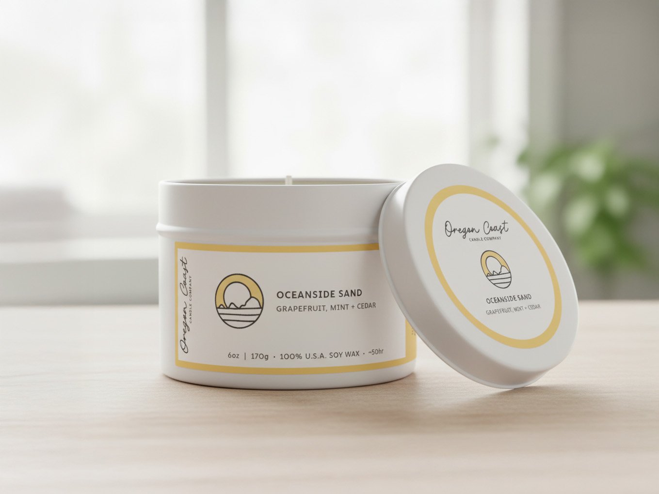 A white candle jar with a matching lid, labeled "Oceanside Sand" with scents grapefruit, mint, and cedar, sitting on a light-colored wooden surface with a blurred window and green plant in the background.