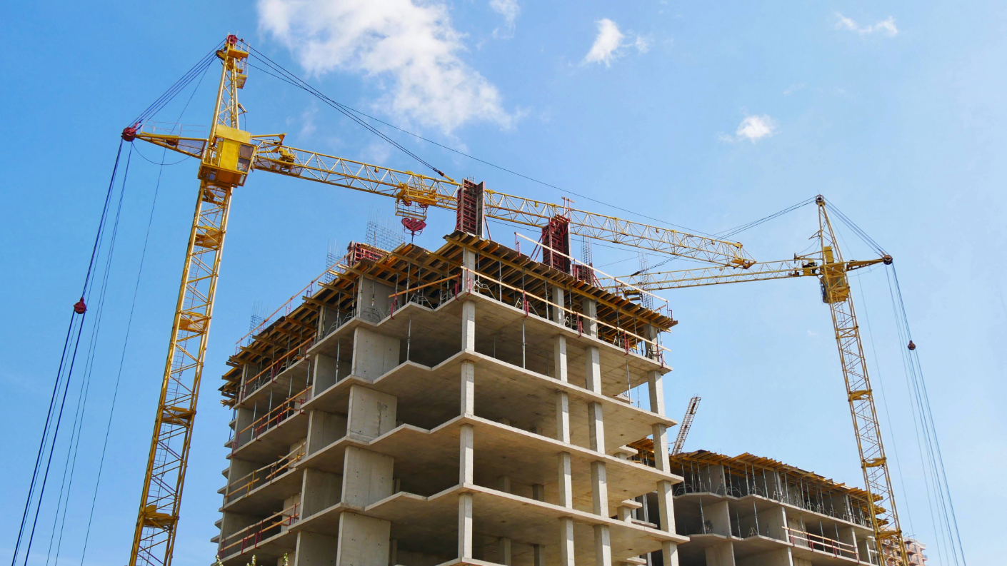 Construction site with a multi-story concrete building under construction, two yellow cranes overhead, and a partly cloudy sky.