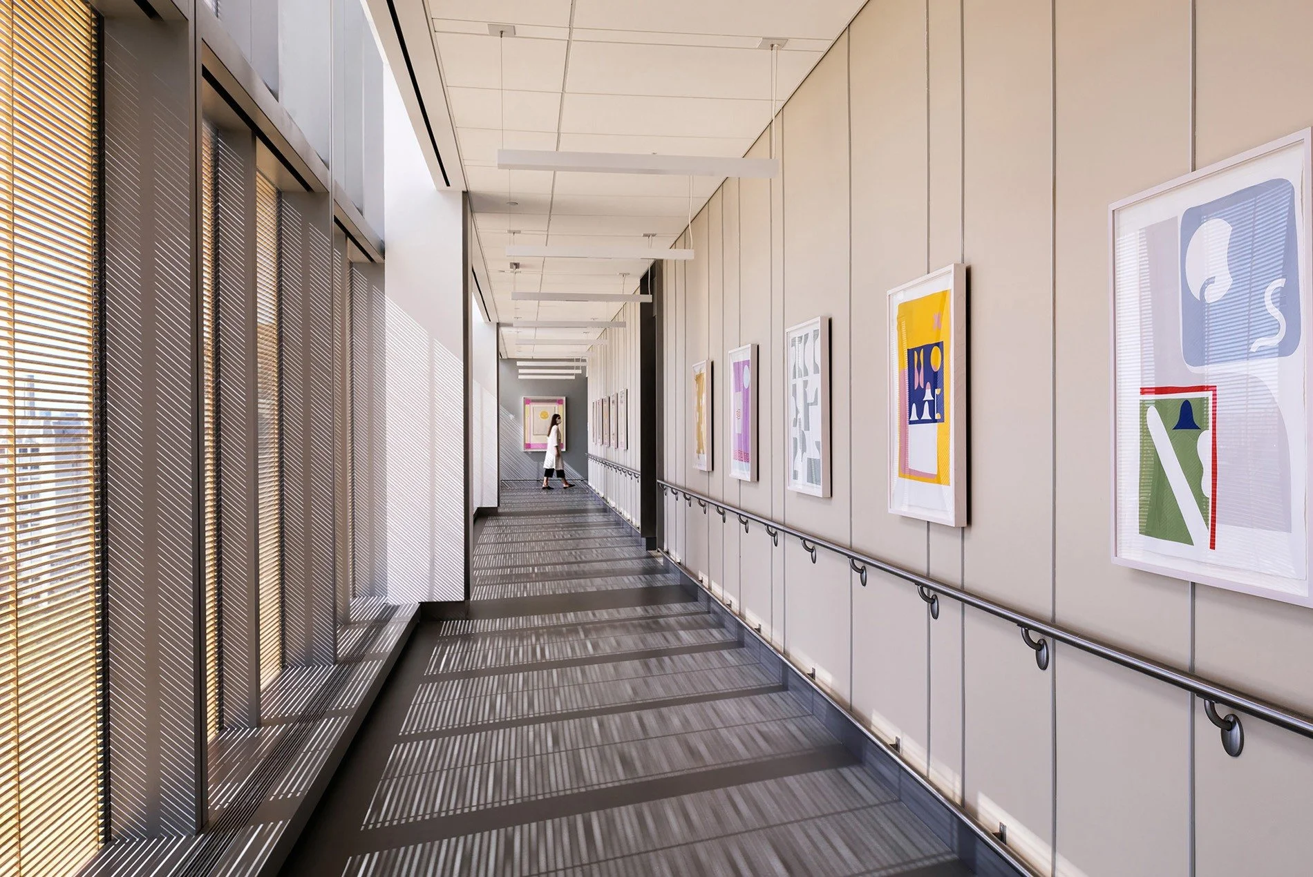 An indoor art gallery hallway with framed abstract artwork on the wall, large windows with blinds casting striped shadows, and a woman in white walking in the distance.