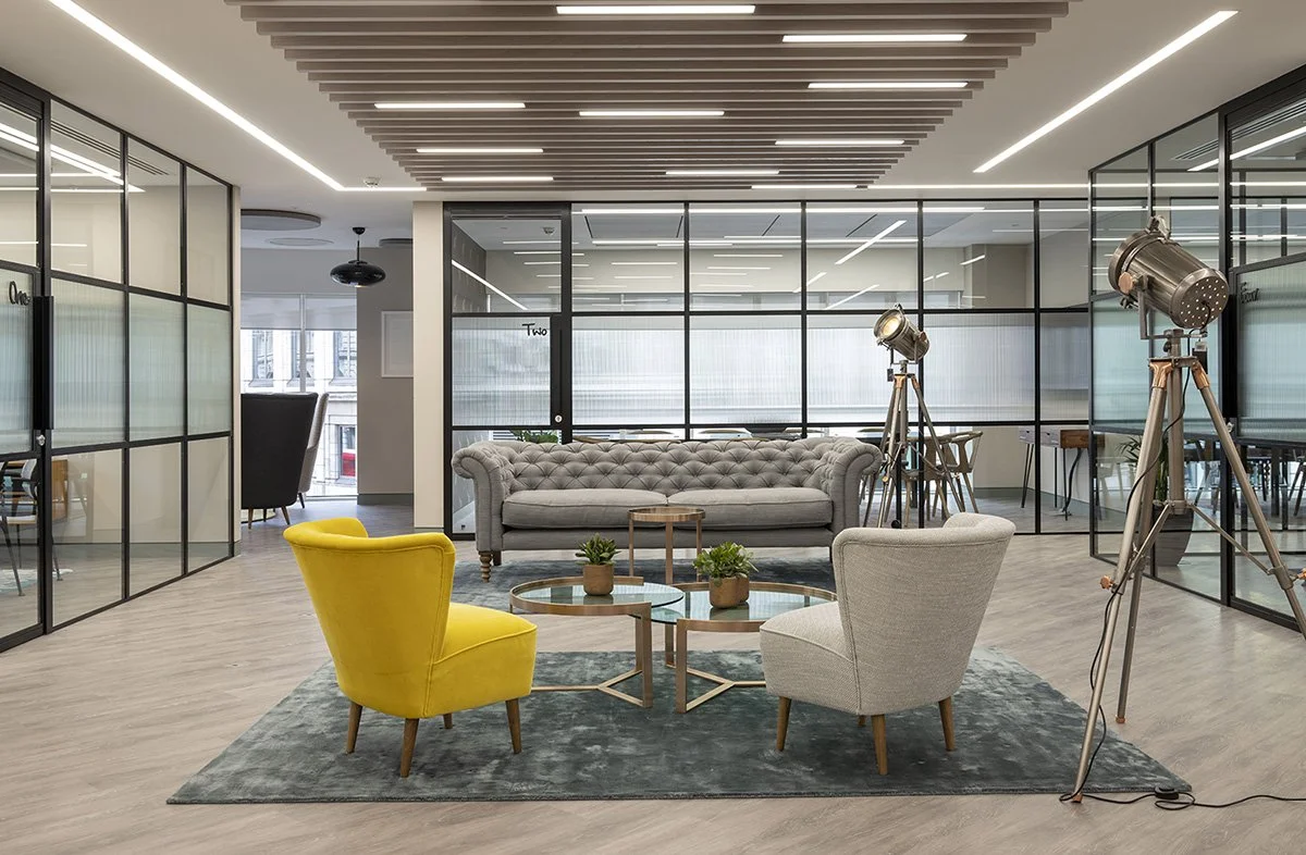 Modern office lounge area with a gray tufted sofa, a yellow armchair, a white armchair, and two vintage film studio lights on tripods. Glass walls divide the space, and there are small potted plants on glass tables.