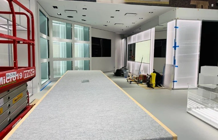 Interior of a commercial space under construction, with a gray carpeted floor, large glass windows, and equipment including scaffolding and tools.