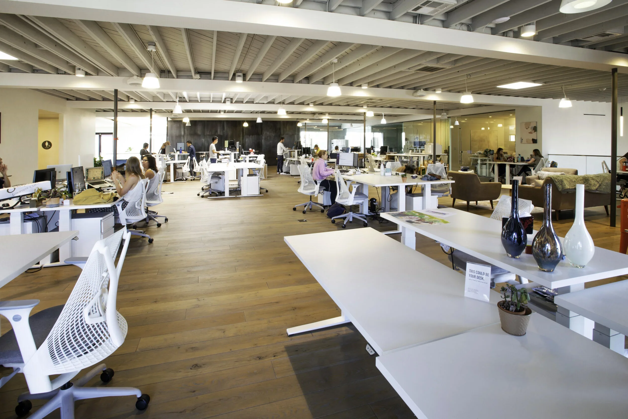 Open-plan modern office with employees working at desks, some seated and some standing, with a mix of white office chairs, couches, and decorative vases, illuminated by ceiling lights.