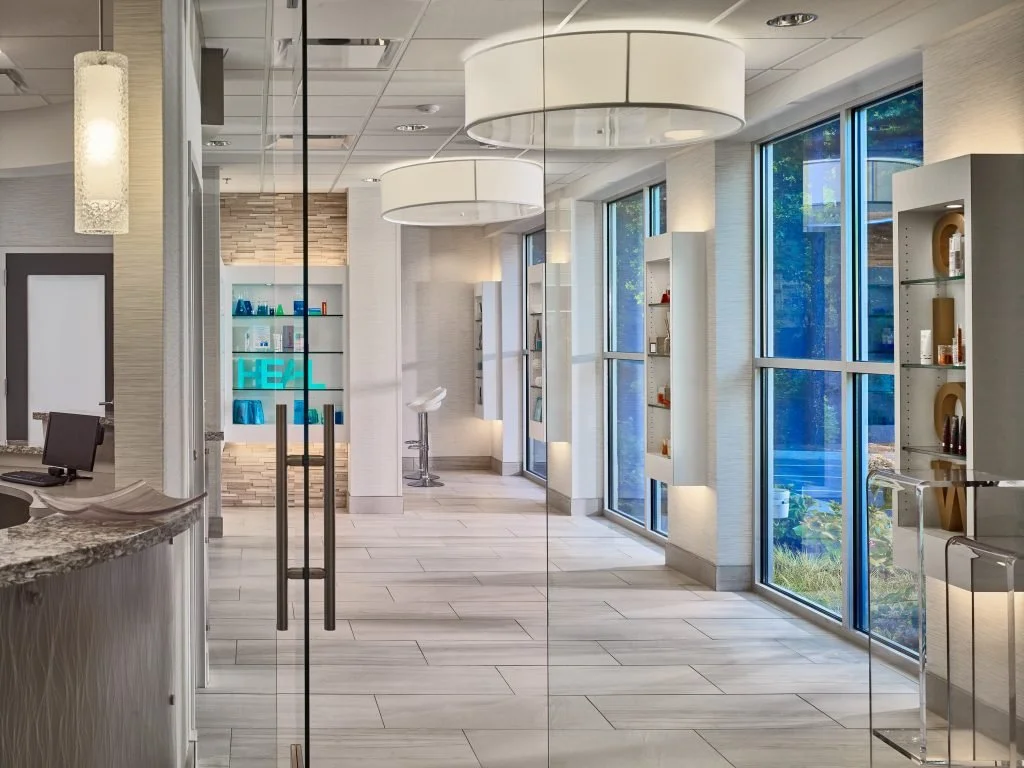 Modern, brightly lit hotel lobby with large windows, white walls, decorative shelves, and minimalist decor.