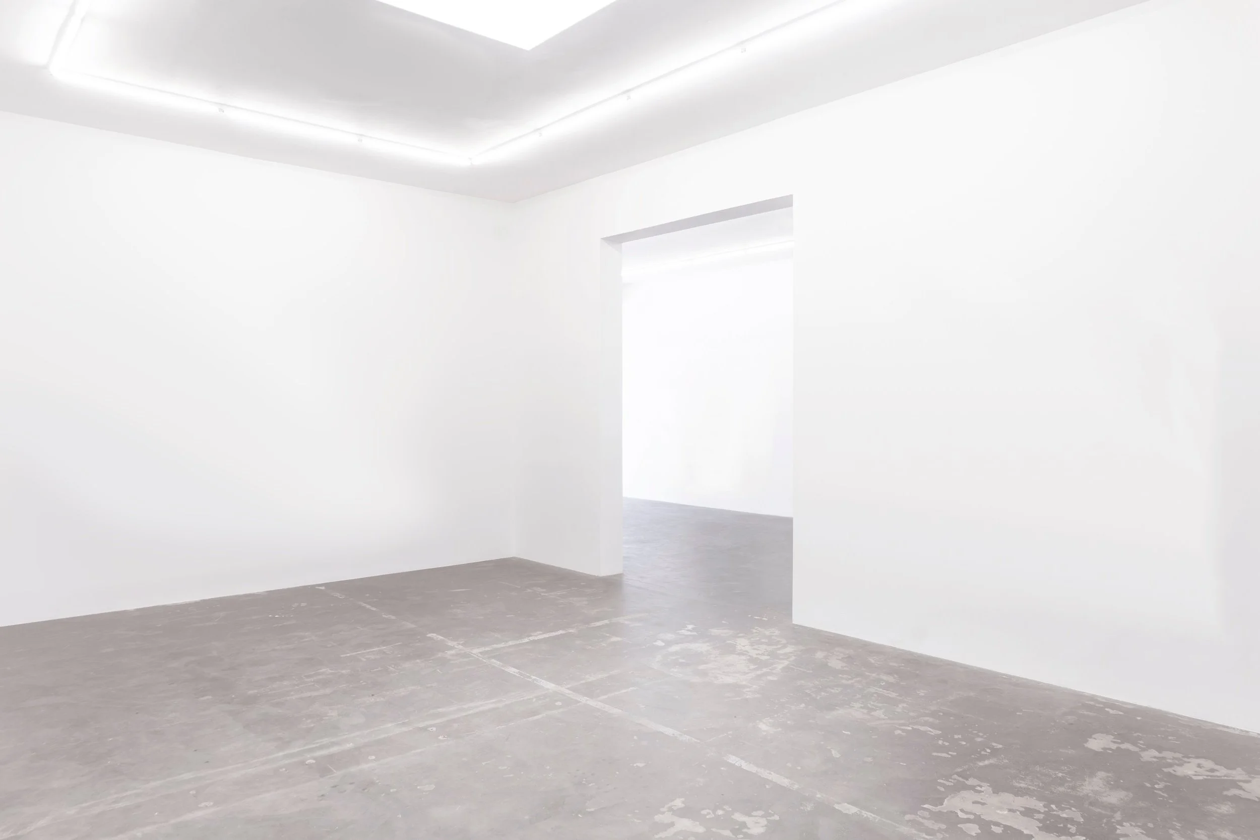 Empty white gallery room with clean walls, concrete floor, and a doorway leading to another bright room.