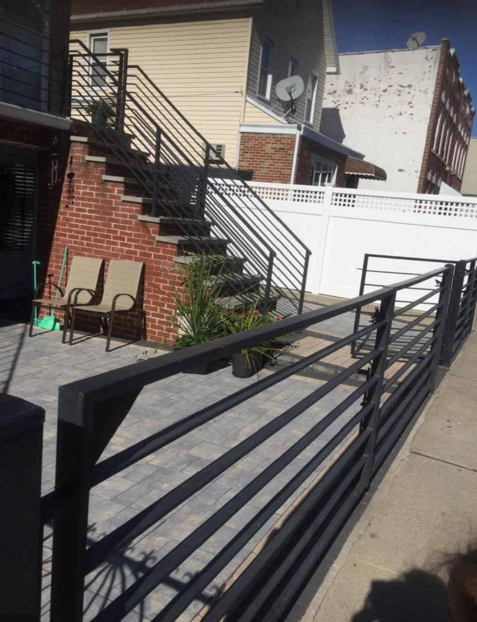 A backyard patio with a brick and siding house, black metal staircase, outdoor seating, potted plants, and a wooden fence, under sunny skies.