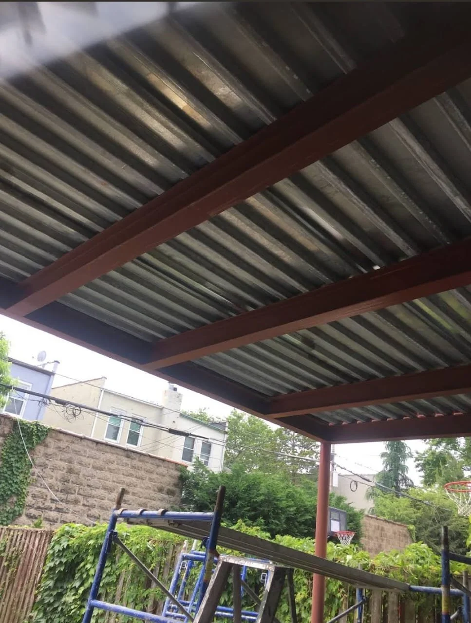 Backyard patio with a metal roof supported by red painted wooden beams, a blue ladder, and a small basketball hoop hanging on the fence, surrounded by trees and residential buildings.