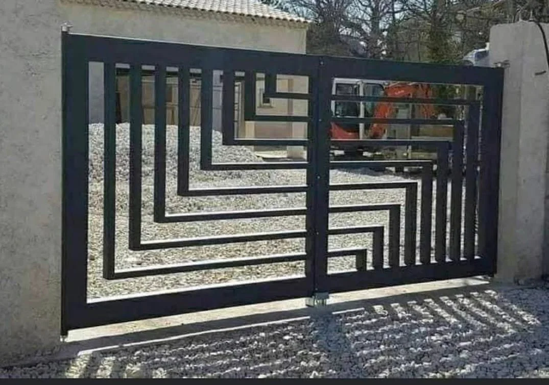 A metal gate with a geometric, maze-like pattern, casting shadows on the gravel ground in front of it.