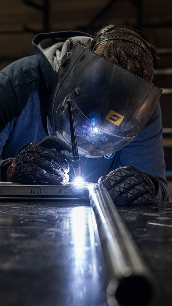 Welder wearing a helmet and gloves welding metal pipe in a workshop.