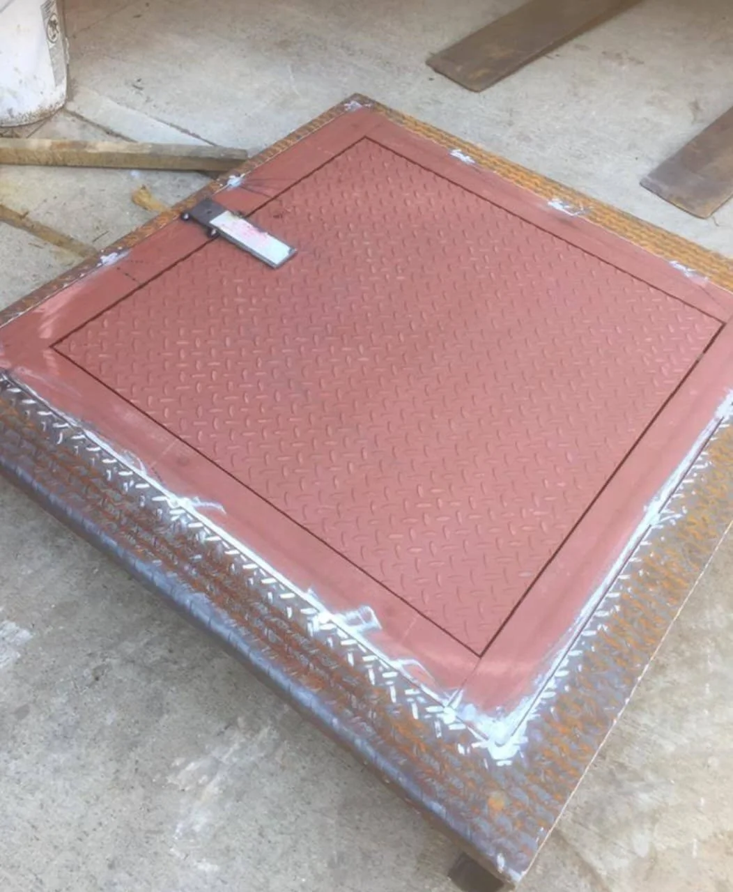A square metal hatch with a textured surface, painted red, surrounded by a rusted metal frame, on a concrete floor. Construction tools and materials are nearby.