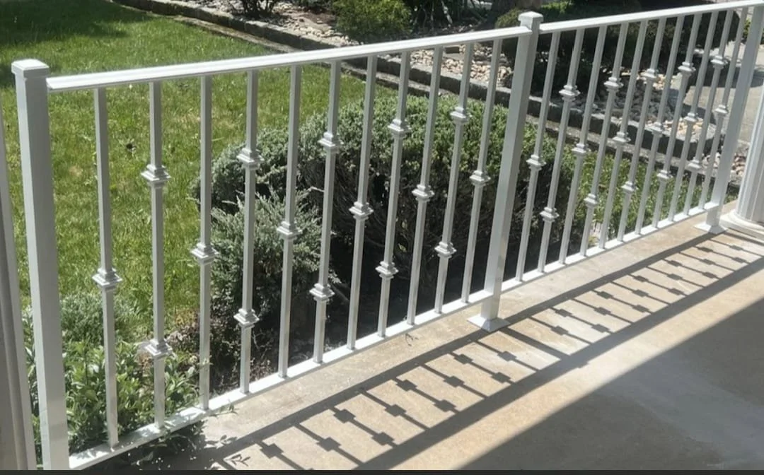 A white metal porch railing casting shadows on the concrete floor, overlooking a garden with green grass and bushes.