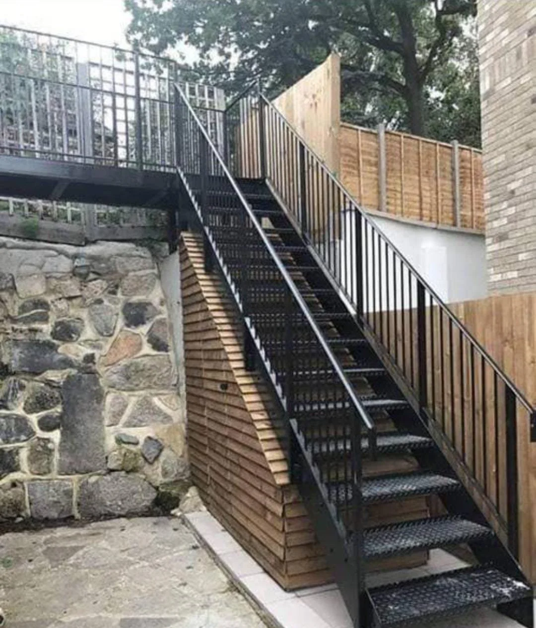 Black metal outdoor staircase with handrails, leading up to an elevated deck with a black railing, adjacent to a stone wall and wooden fencing.