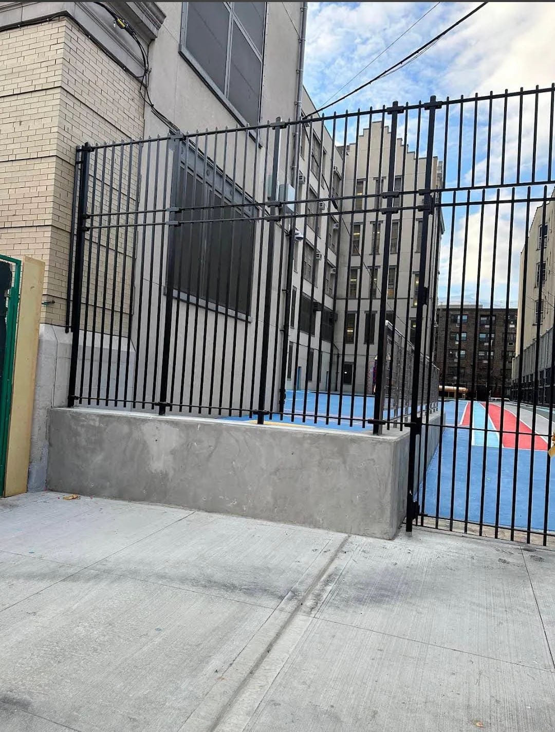 Fenced sports court with blue and red surfaces behind a black metal fence, located in an urban area with multi-story buildings.